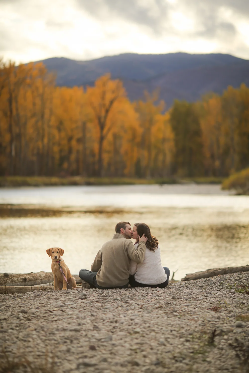 Couple kissing with puppy sitting next to Montana river during fall engagement portrait