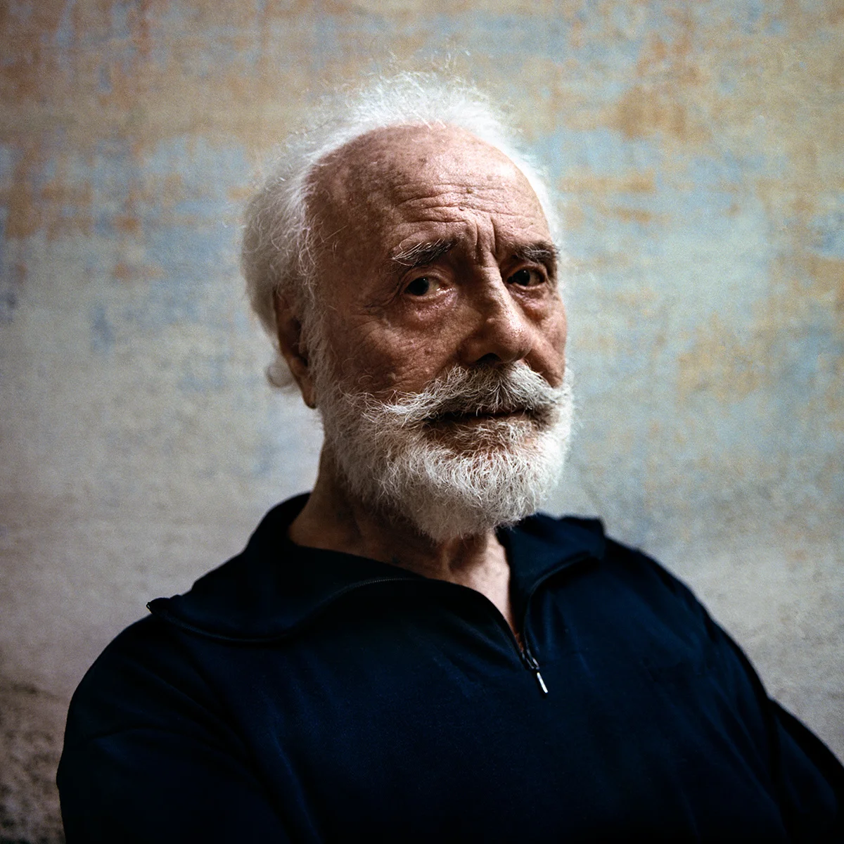 Pino Lella is one of those people with an aura that ends up influencing their life. I felt it when I met him. It becomes clear when you learn about his life. I didn’t know anything about Pino until I read the novel based on his life. I read it fast, it consumed me. When I finished the last page I was sitting at the Denver airport weeping. I knew right there and then that I had to meet this man. It was one of those things that would keep me up at night until I got it out of my system by going and photographing it. I did have a connection that would allow me to get my foot in the door and I emailed him right there and then. I was going to be in Austria, about 4 hours from where Pino currently lives, about a week and a half later and I could drive to meet him. It was a long shot but I had to try. Over the next ten days I went to Romania and then Austria. All the while trying to get a commitment out of Pino’s daughter for me to come and take a portrait of Pino. The family was guarding him not only due to his age but also because of legal concerns since a movie adaptation of Pino’s story was underway. The day before the last available window I finally secured my chance to meet Pino through his wife. The next morning I got in the car and crossed the alps into Italy to meet Pino. Then, just about 20 minutes from his house the road was closed and a police man told me that there was no getting through. I spent about 20 minutes trying to come up with a way around but since the road was running along a large lake none of my attempts were fruitful. I started worrying. If I didn’t figure this out within the next 10 minutes I was going to be late and possibly not able to make it all. I said to myself: No! This is not happening! I am not going to be shut down last second. There has to be a way. So I decided to go through where the police man told me not to. And I made it. Pino’s wife Yvonne welcomed me into the home and after she showed me around a little I started taking a few portraits of her. Then Pino came jogging up the narrow alley and I introduced myself. Quickly we started shooting since I was urged to keep it short. I took a few portraits in their courtyard and then used a beautifully textured wall in the alley with the light coming pretty strongly from one side as a background. I started with my go-to-pose of a very straight-on and emotionless look. The light was a little too much from the side for it so we started playing around a little and I had Pino turn towards the light. There I got two fantastic portraits. One of them reminds me of a classical painting, or a greek god. I knew I got something good there so I cut it short by only shooting one more roll of film there and then switching to a full length shot around the corner in another alley. It was clear that the previous shot was going to be it so we didn’t spend too much time. We chatted for a few minutes, I thanked them excessively for being open to me photographing them and then right before I was going to leave I asked Pino: Tell me about James Dean. And from my left Yvonne goes: You know, he was supposed to be the one driving him that day… Between me first learning about Pino and standing in front of him lay only ten days. There were however 6000 miles between it, gatekeepers, the alps, legal challenges and not to forget an Italian police man who could have made all efforts go puff last second. When I got back in my car and drove along the shore of Lago Maggiore back to Austria I got overwhelmed by a feeling of ridiculous accomplishment. An accomplishment that had defied all odds.