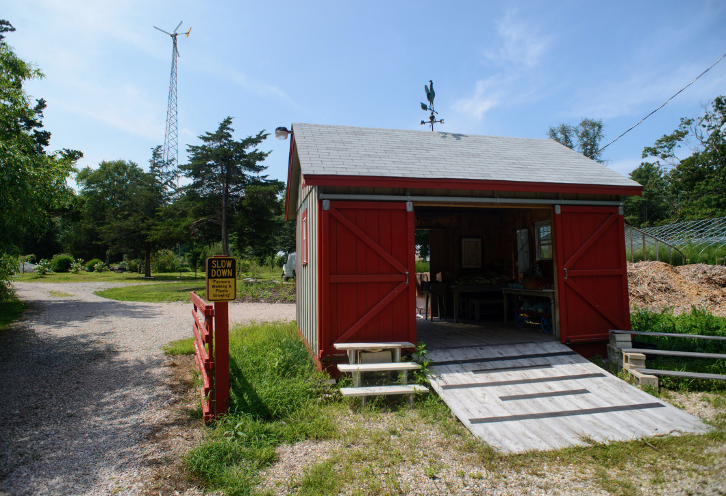 A small red barn with open sliding doors, a ramp leading inside, and a small staircase outside. Surrounding greenery, trees, and a gravel driveway with a yellow sign that reads "Slow Down" in the foreground.