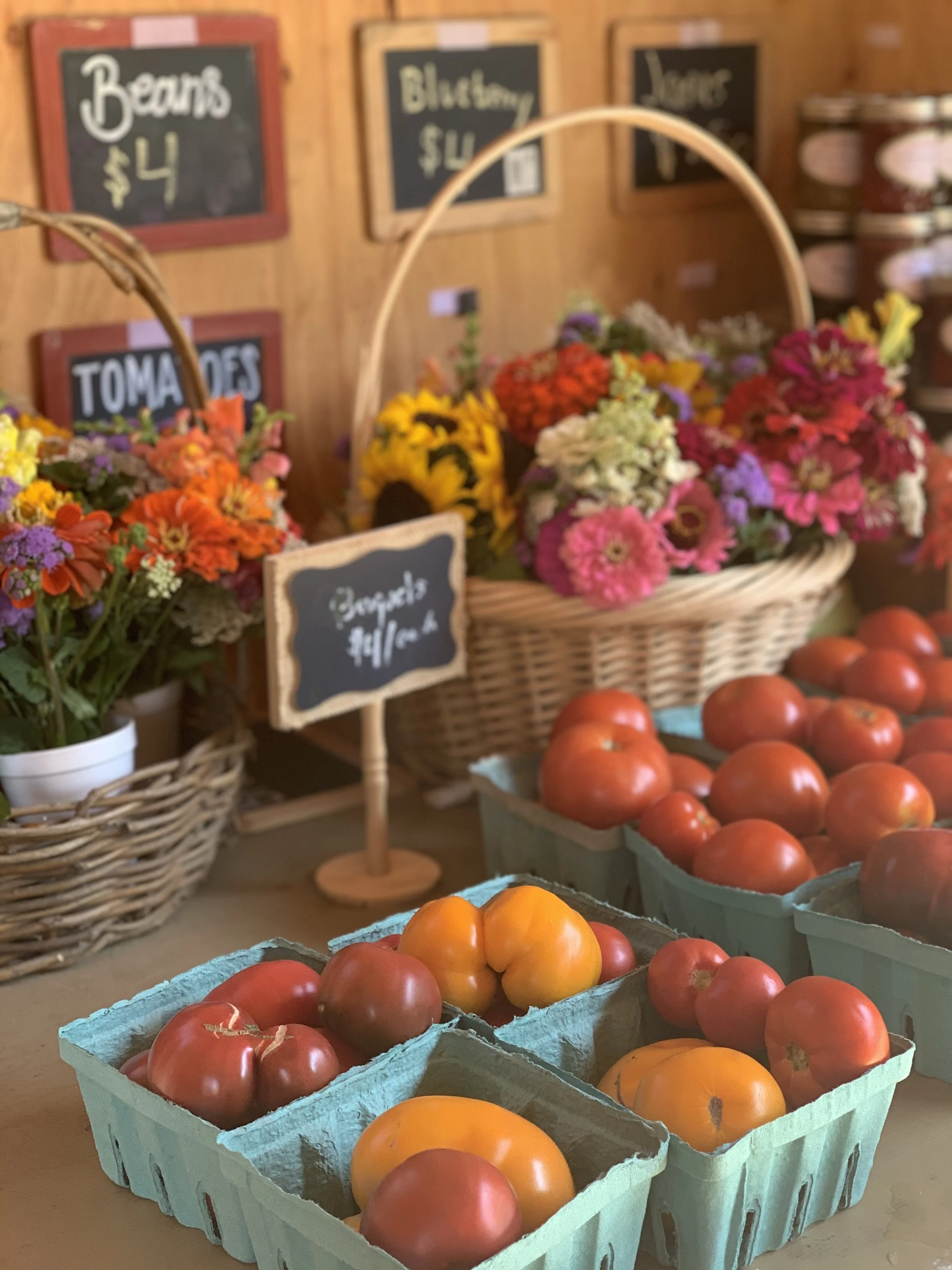 Fresh tomatoes in green baskets on a wooden table with flowers in baskets in the background at a farmers' market.