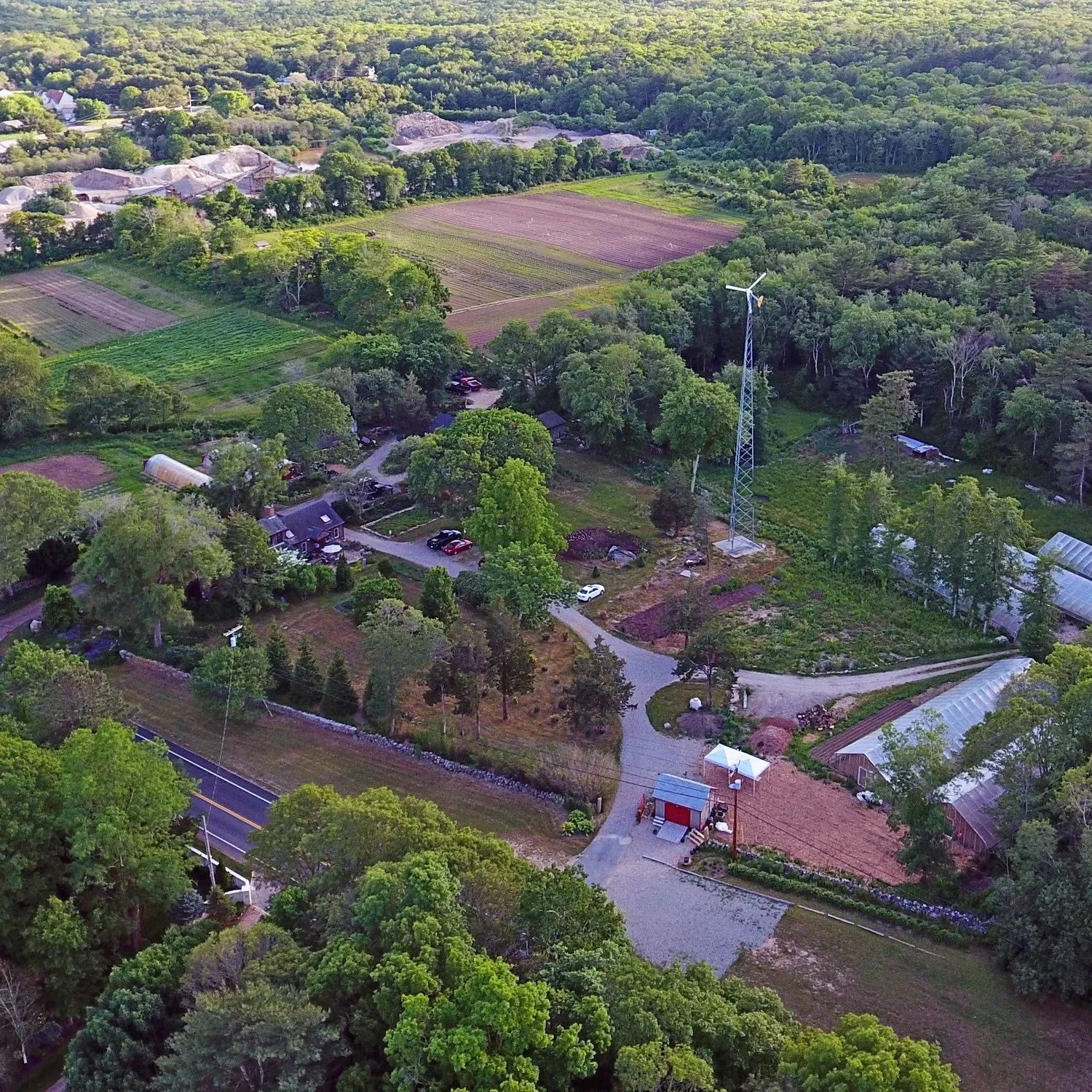 Aerial view of a rural landscape with a mix of farmland, greenhouses, houses, trees, and a wind turbine in the background.