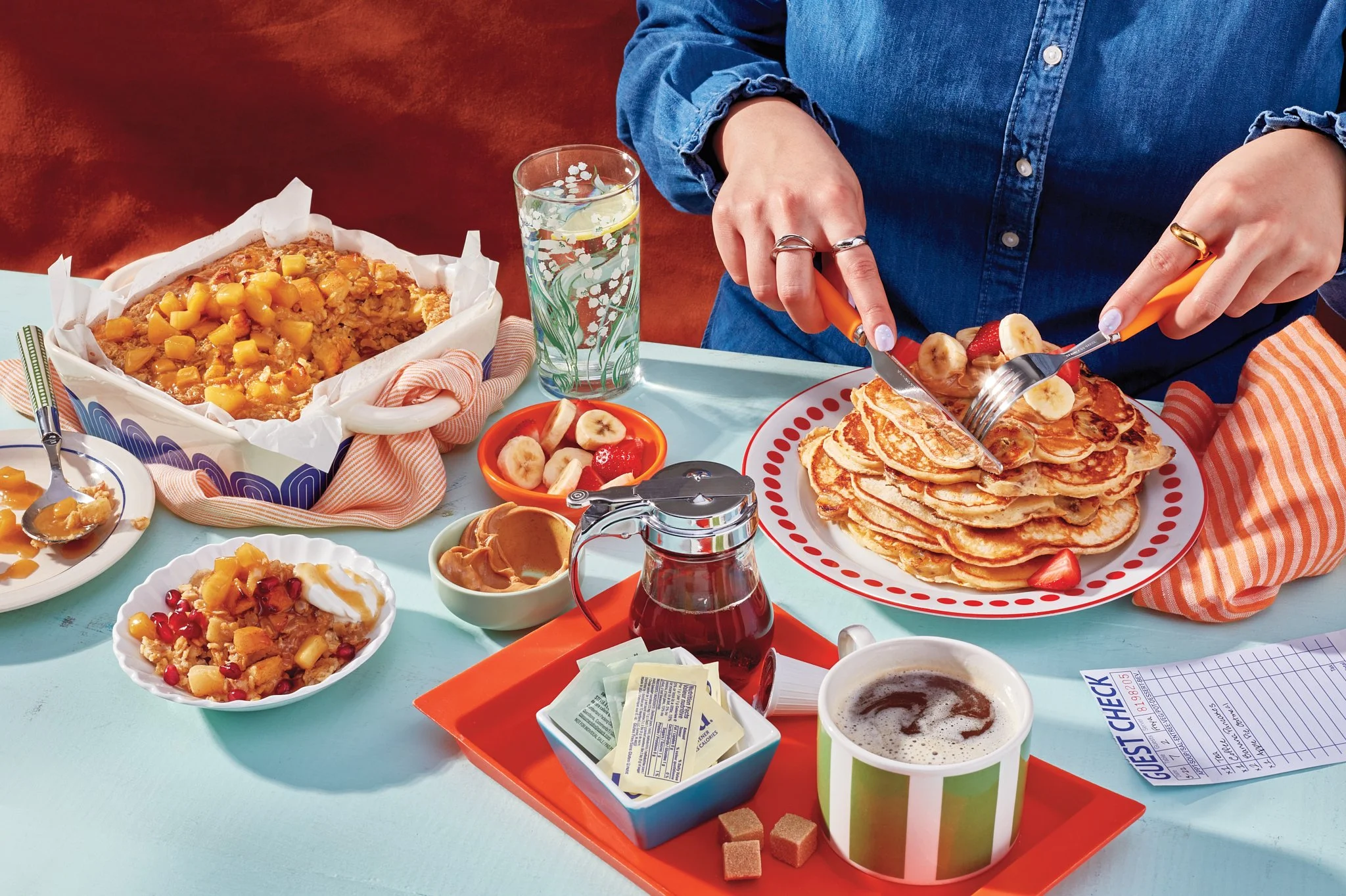 A breakfast spread on a table with a person in a blue denim shirt, cutting pancakes with banana and strawberry slices. The table has various bowls of fruit, syrup, a glass of water with lemon, and a cup of coffee.