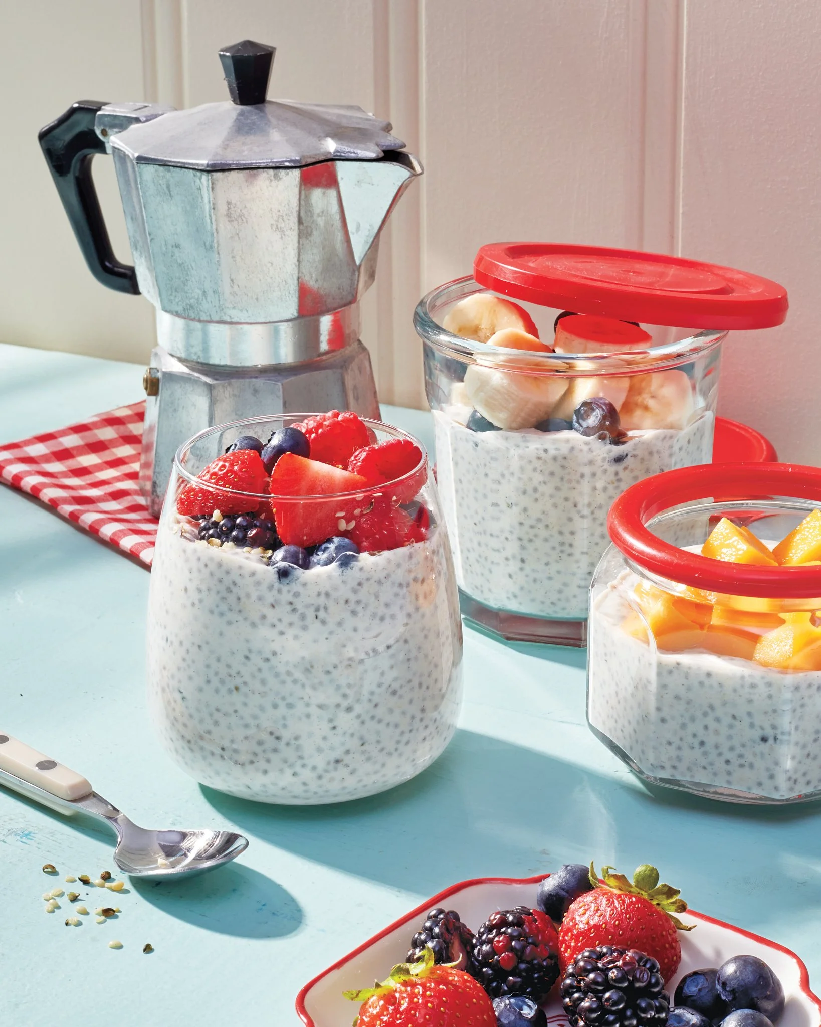 Various containers of mixed berries, banana slices, and a moka pot on a light blue surface with a red and white checkered cloth.