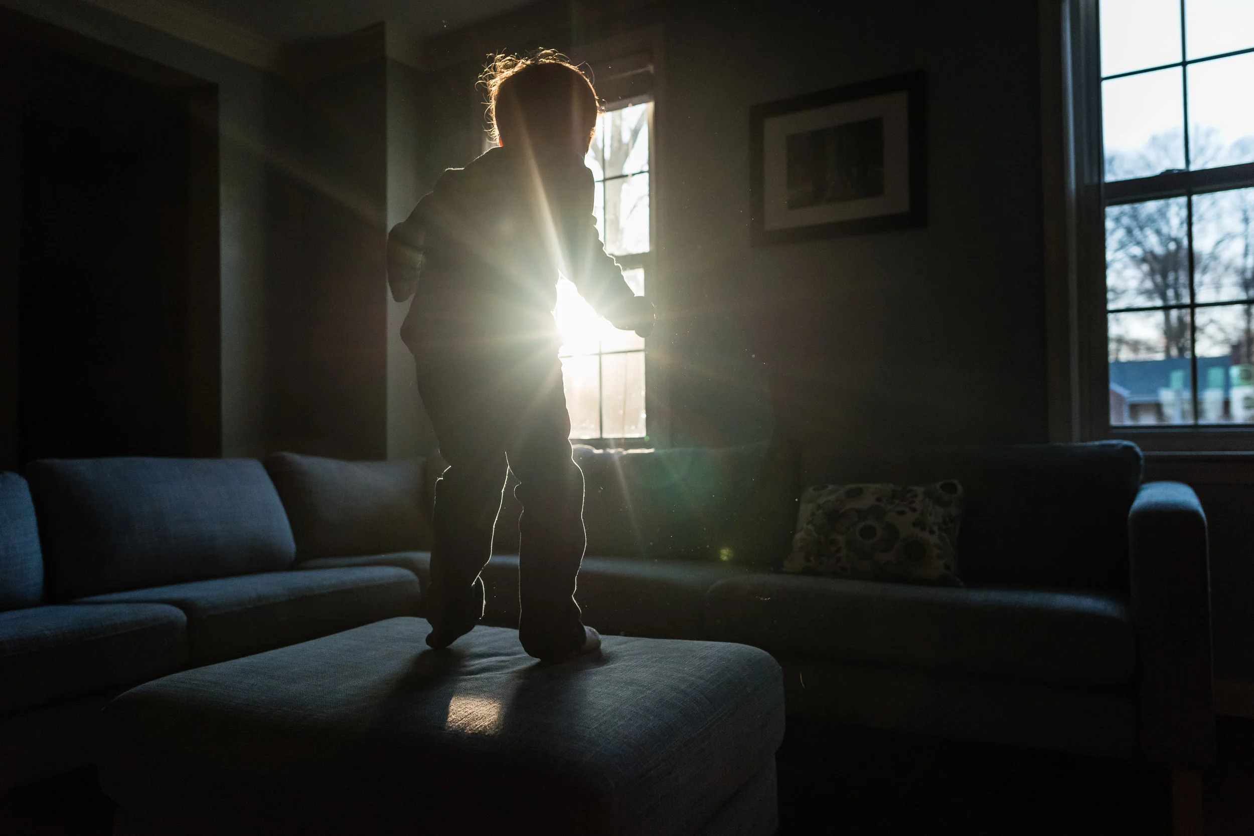Boy jumping on ottoman with sun flare in Northern Virginia by Family Photographer Nicole Sanchez