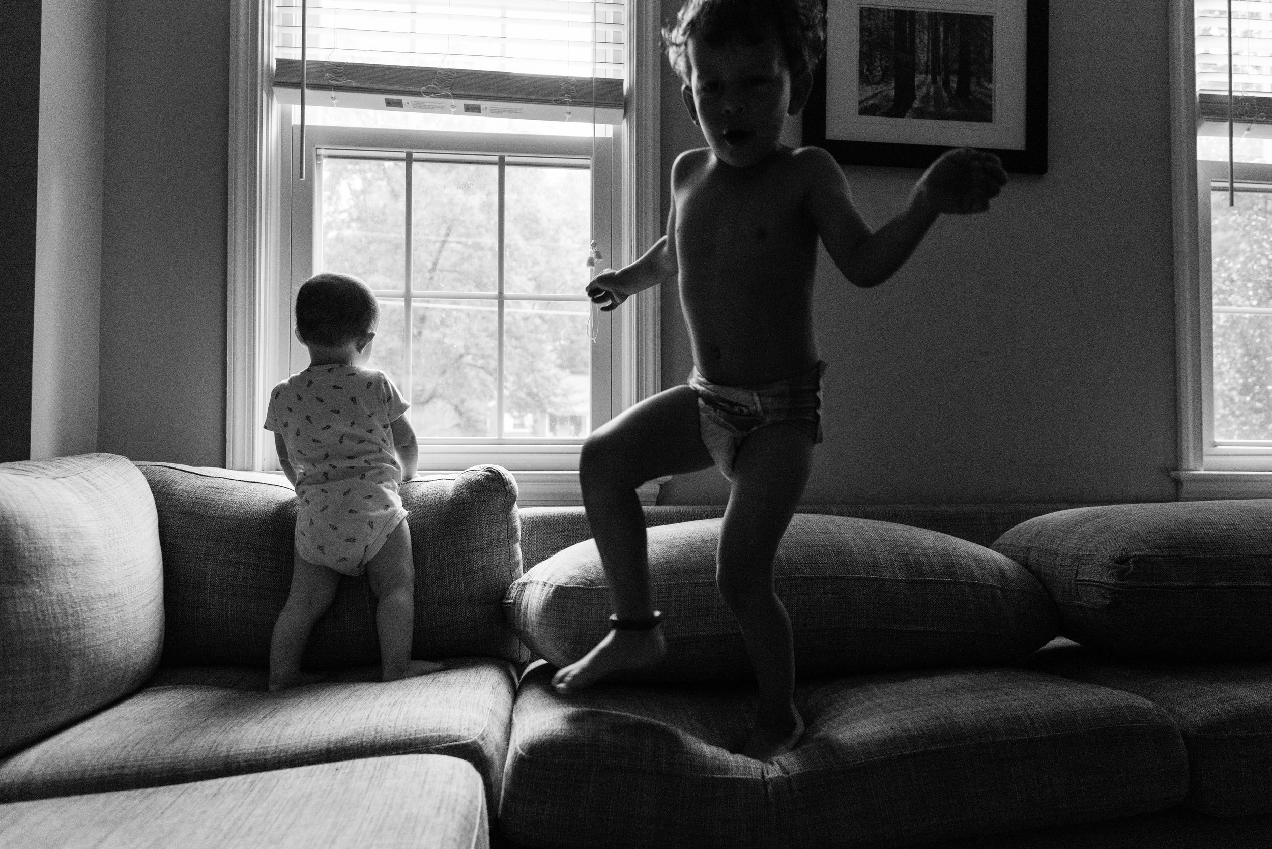 baby looking out window while brother jumps on couch by northern virginia family photographer nicole sanchez