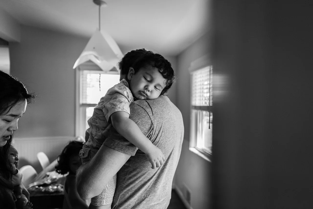 Toddler boy sleeping on Dad’s shoulder while Mom takes shoes off by Northern Virginia Family Photographer Nicole Sanchez