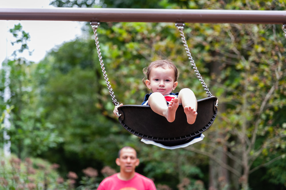 Toddler swinging on Northern Virginia playground with dad in background by Family Photographer Nicole Sanchez