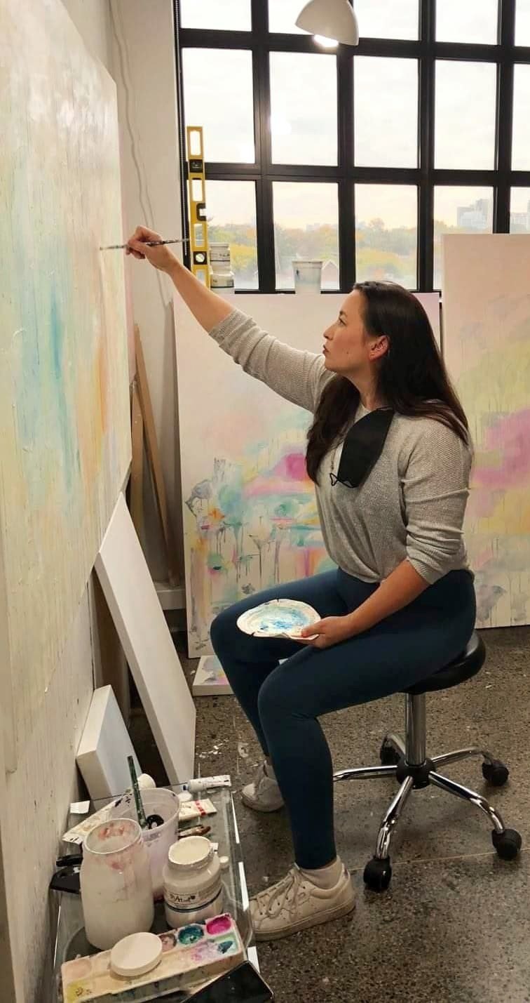   Linds Miyo sits on a stool inside her studio, a palette in one hand and a brush in the other, painting on a canvas mounted to the wall. Below her are various paint jars, brushes and palettes. Behind Linds is a large window, resting on which are mor