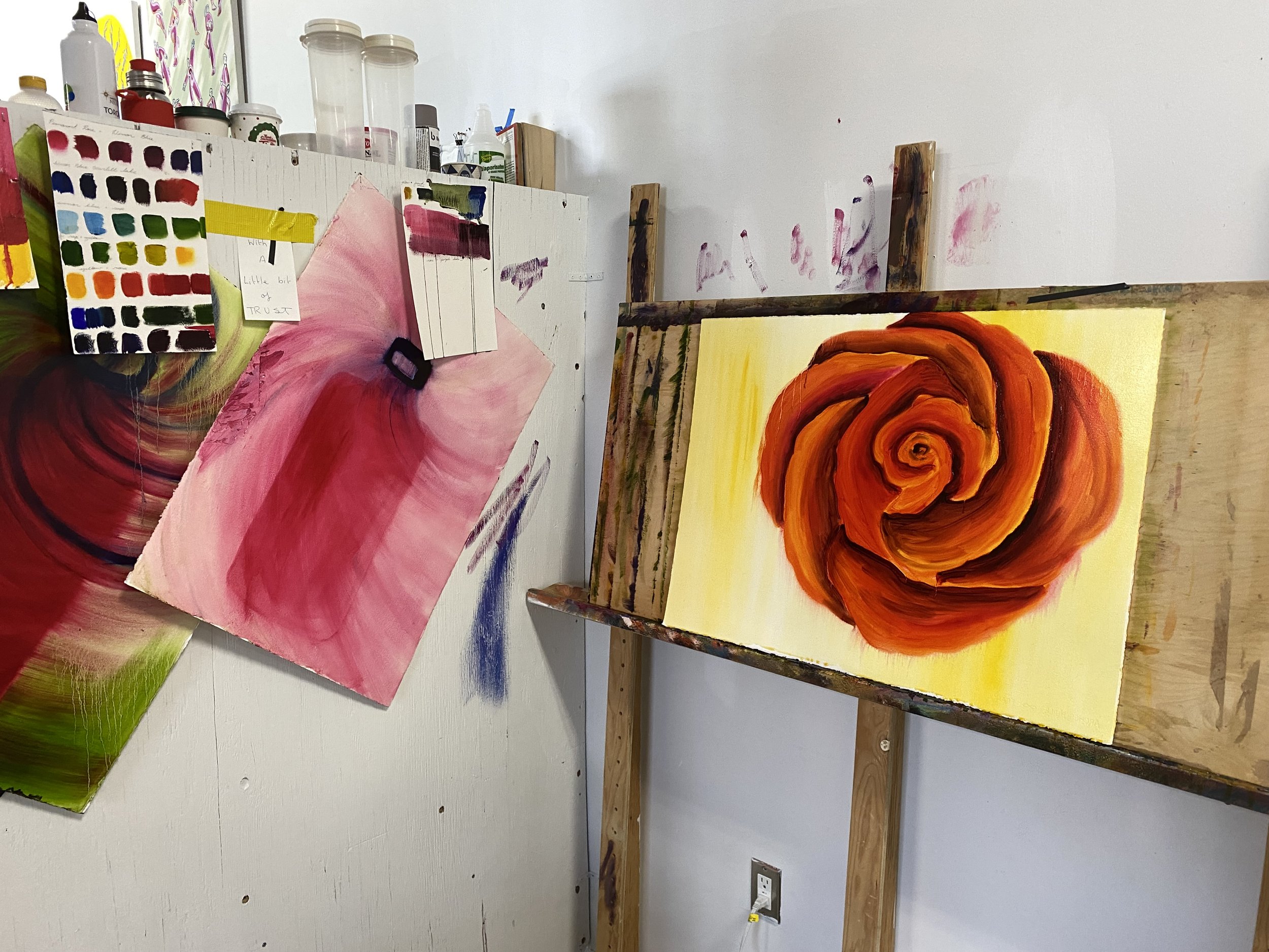   Kate Gorman’s studio, featuring a work in progress. To the left corner of the wall, paint swatches, colour tests and hand written notes are taped to the wall, amidst larger abstract paintings on paper of vivid pinks, reds, and greens. On the right 