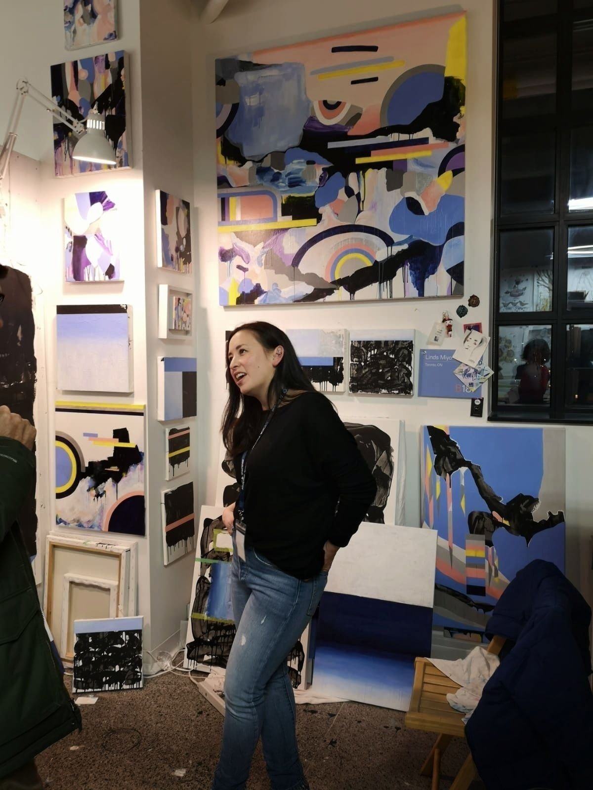   Linds Miyo standing in her studio, surrounded by her paintings. She is talking to someone who is seen partially out of frame of the photo.   