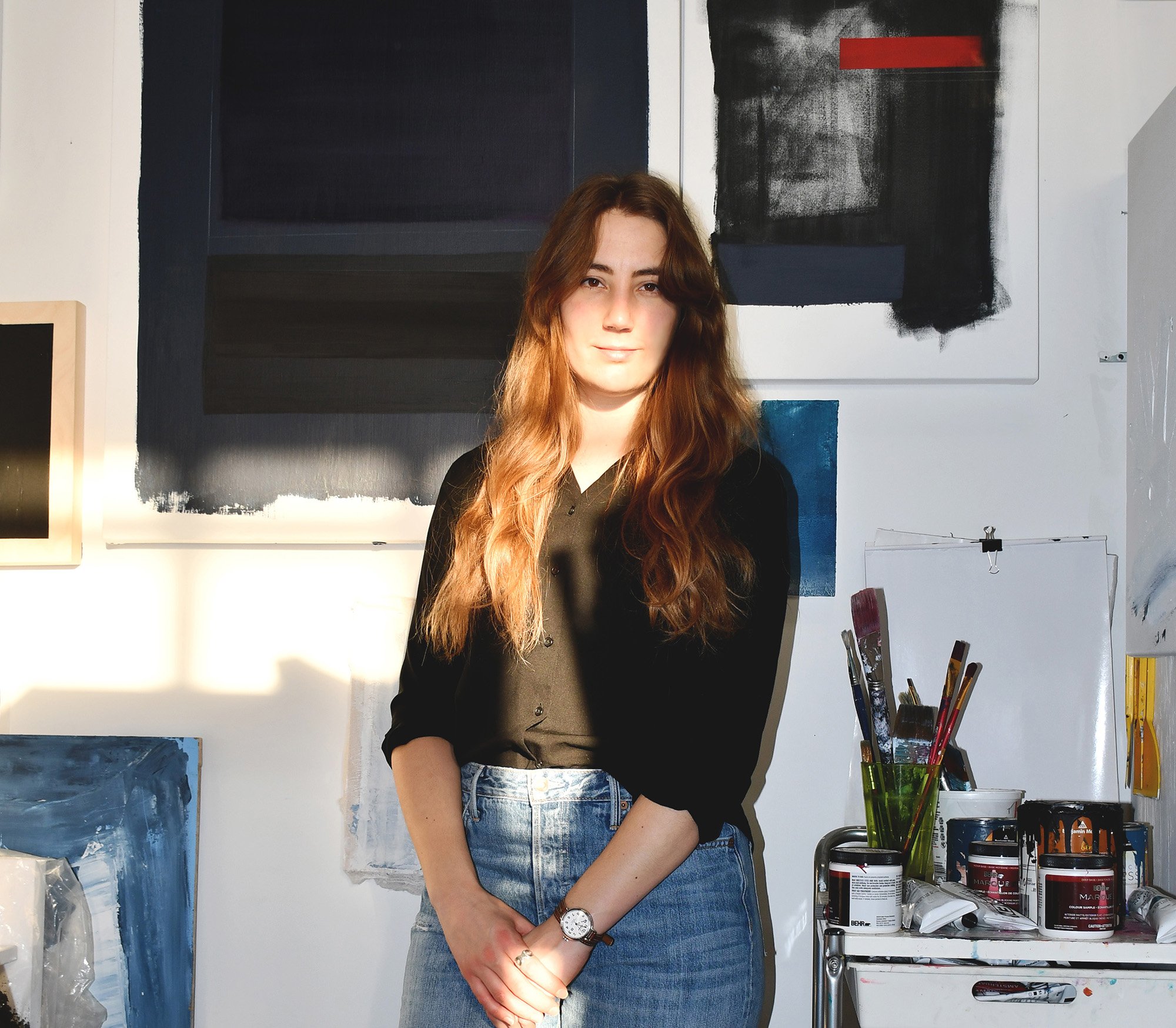   Mel Hayes is standing in her studio, she is wearing blue jeans, a black shirt, and has long brown hair. Behind her are paintings, mounted to the walls. To her left is a table filled with paints and brushes.&nbsp;  