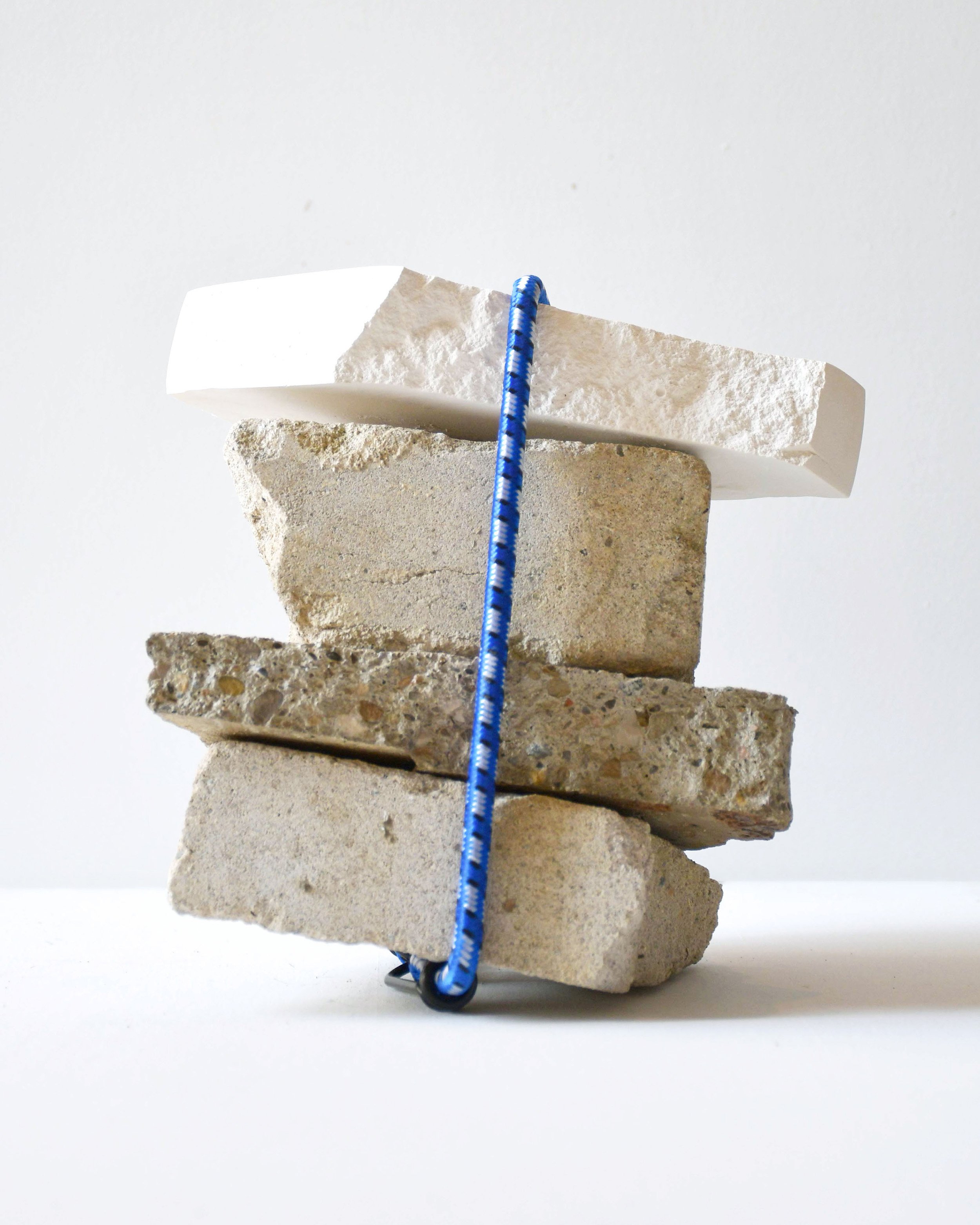   An image of Mel Hayes’ sculpture, Hold Me Time. Four rectangular blocks of white concrete and stone are held together by a blue bungee cord on a white surface against a white background.&nbsp;  