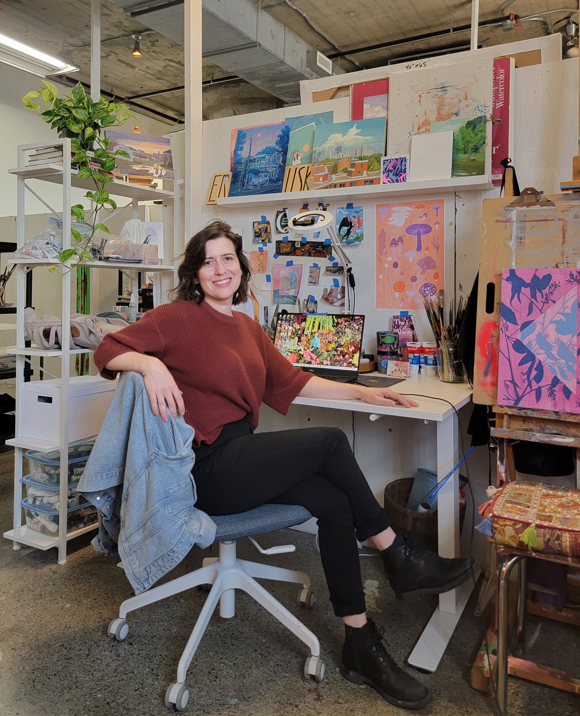   Erin McCluskey sits on an office chair inside her studio. On the desk behind her is her laptop, brushes and paint. Paintings and sketches are taped with blue tape onto the wall behind the desk. Above is a shelf containing more paintings and a green