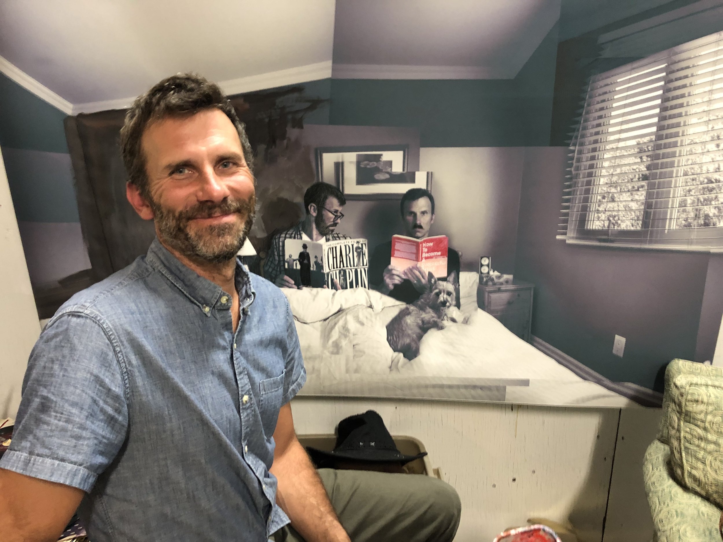   Jason Bomers sits in his studio, smiling. His hair and beard is salt and pepper. He wears a blue button-up shirt and green pants. Behind him is an image of two men in bed, with a small dog, one is reading “The complete films of Charlie Chaplin,” th