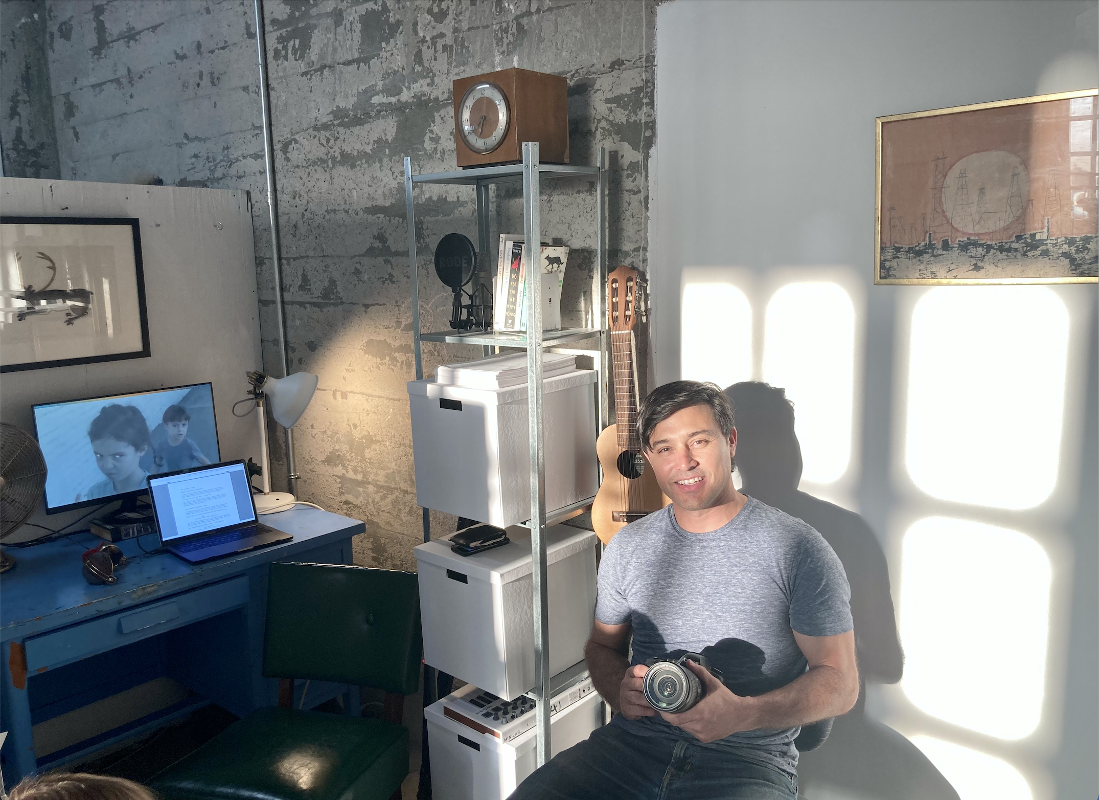   An image of Yani Gellman in his studio. He’s wearing a grey t-shirt and jeans and is holding a camera. To his left is a blue desk with two computer screens open, one to a word document, the other to a blue image of two children reminiscent of a fil
