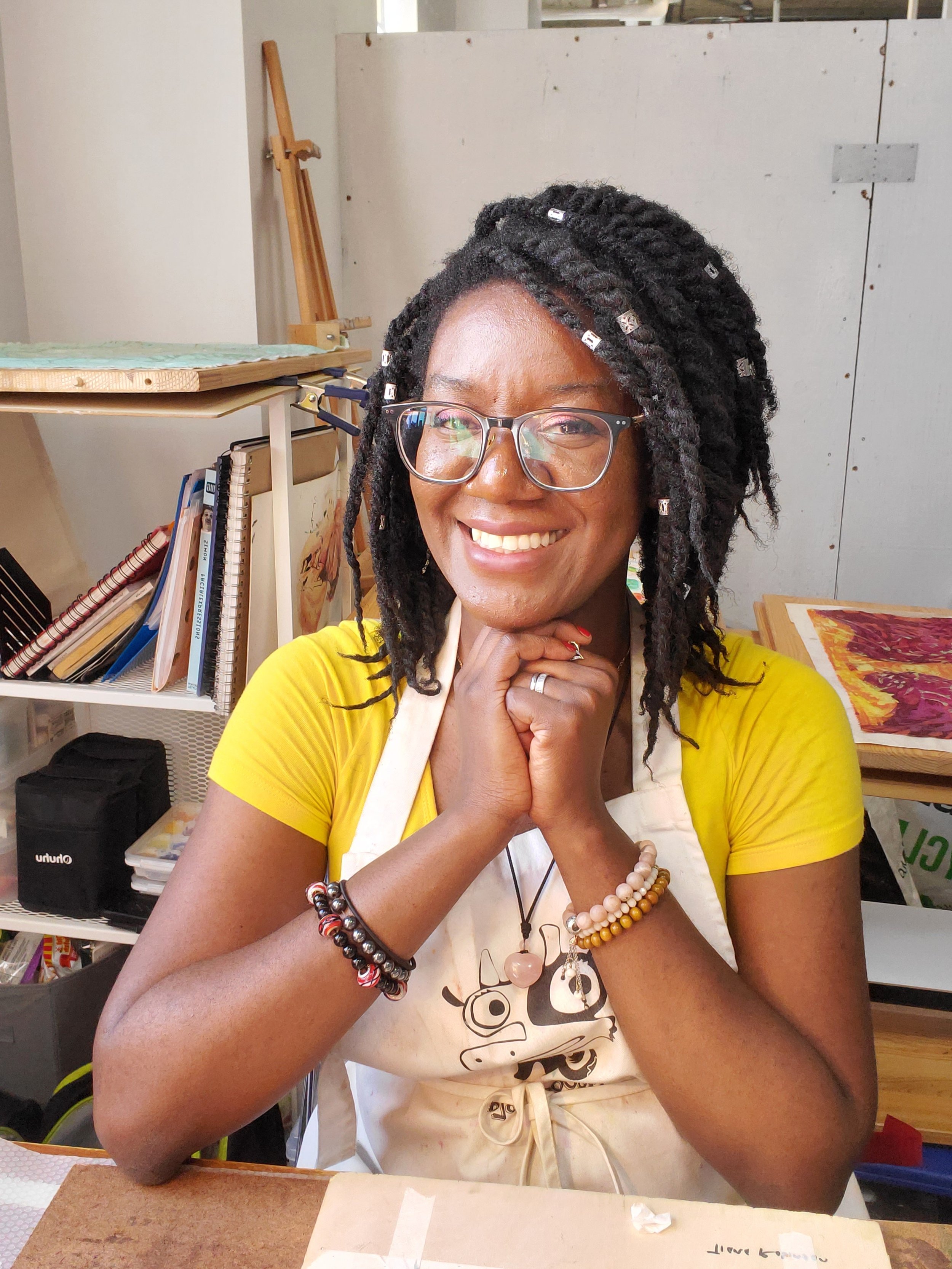   Tiana Robinson sits inside her studio. She wears a bright yellow shirt and has silver beads in her hair. She wears glasses and many bracelets and rings. Her elbows are on her desk, her hands folded under her chin. Behind her are shelves filled with