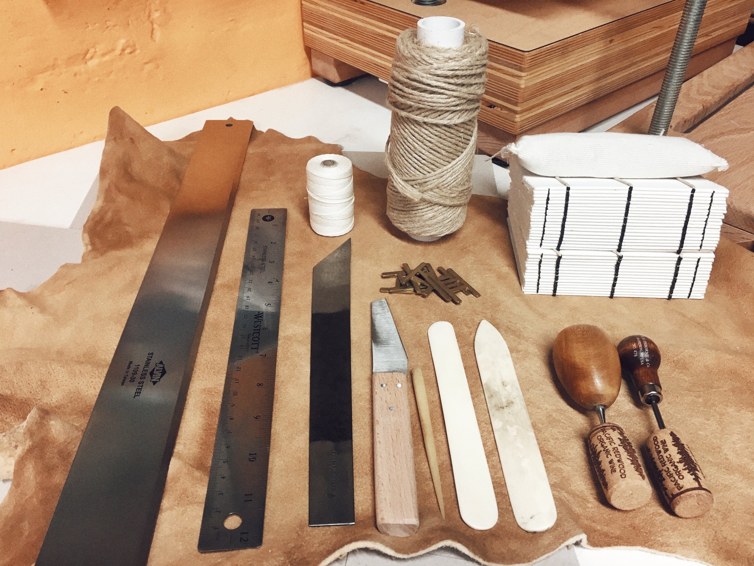 A selection of bookbinding tools. Clockwise from the top right : Blank text blocks, twine and linen thread, tear bar, ruler, English paring knife, leather knife, several bone folds and awls, and sewing keys in the middle. You can see hand-tanned leather as a backdrop and a bit of my press and sewing frame in the background.