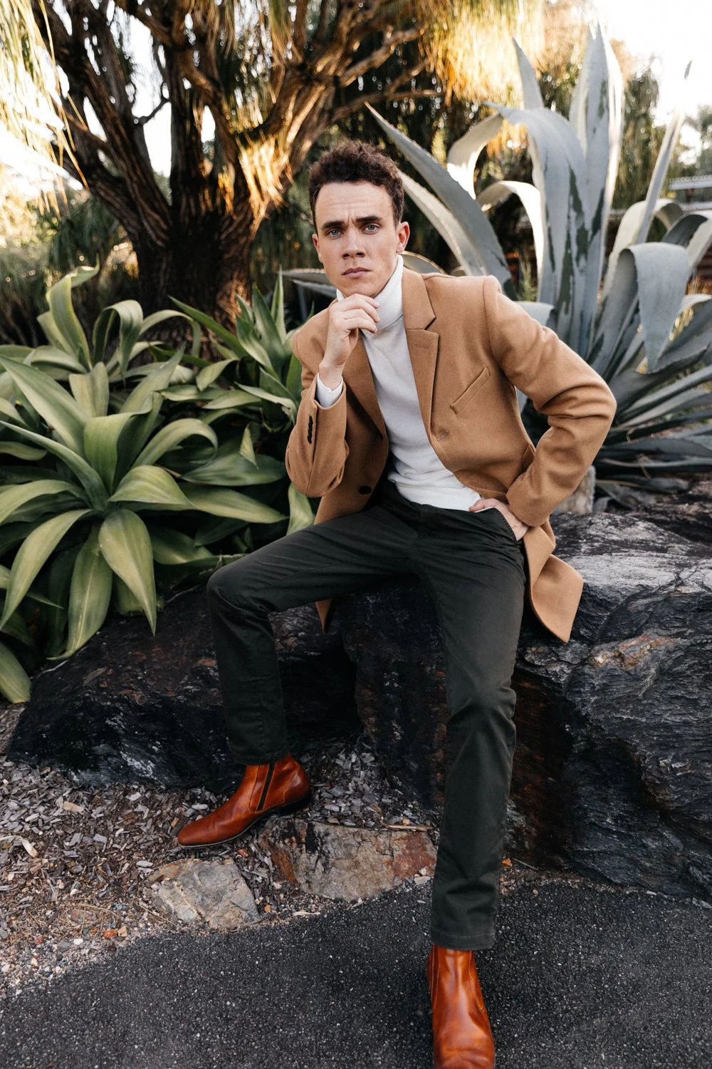 A man in a tan coat, white turtleneck, black pants, and brown boots is sitting on a large rock outdoors surrounded by green plants and trees, posing thoughtfully with his hand on his chin.
