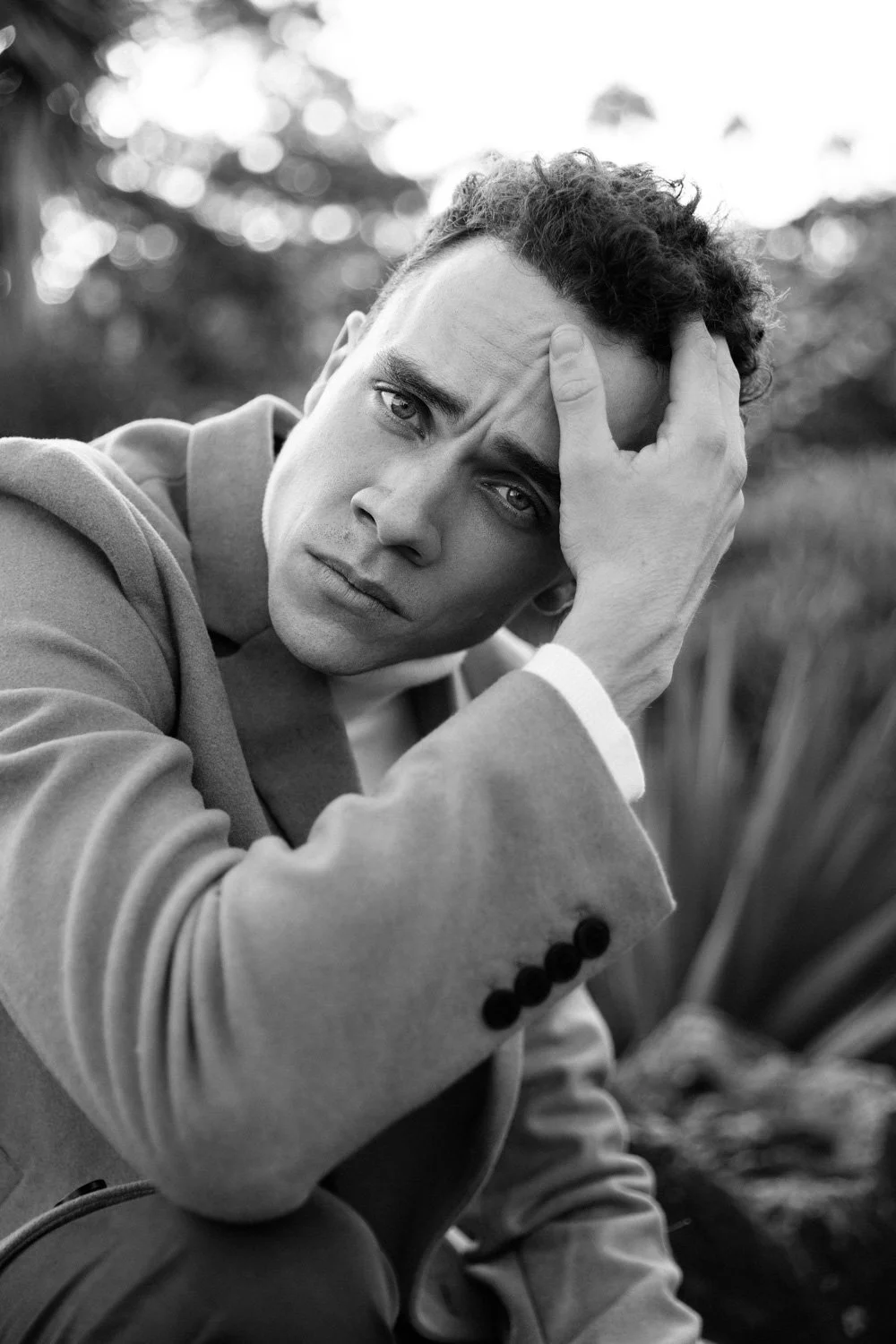 A black-and-white portrait of a young man with curly hair, wearing a blazer, sitting outdoors with a contemplative expression, hand on his forehead, blurred natural background.
