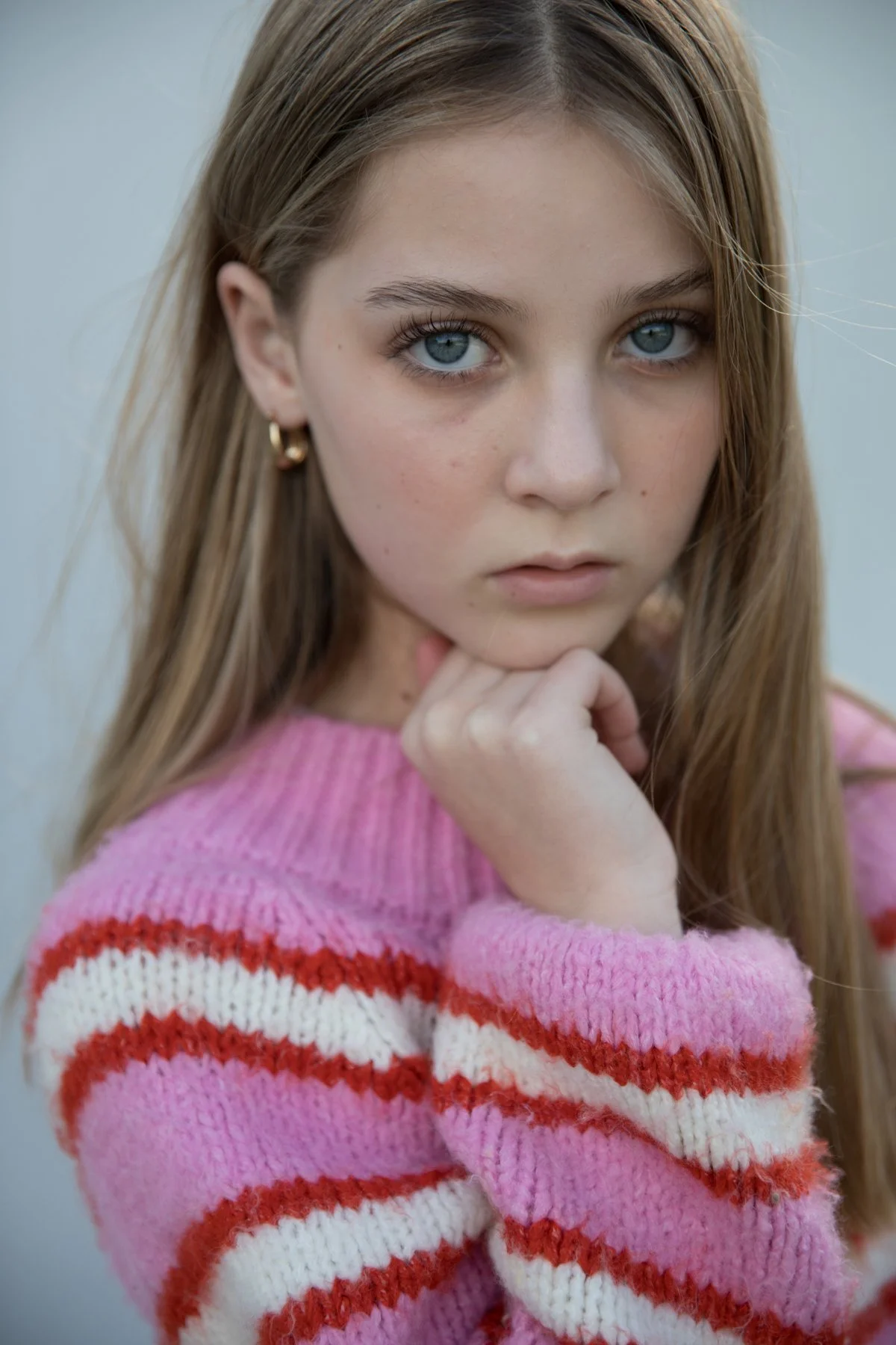A young girl with long light brown hair and blue eyes wearing a pink sweater with red and white stripes, resting her chin on her hand and looking into the camera.