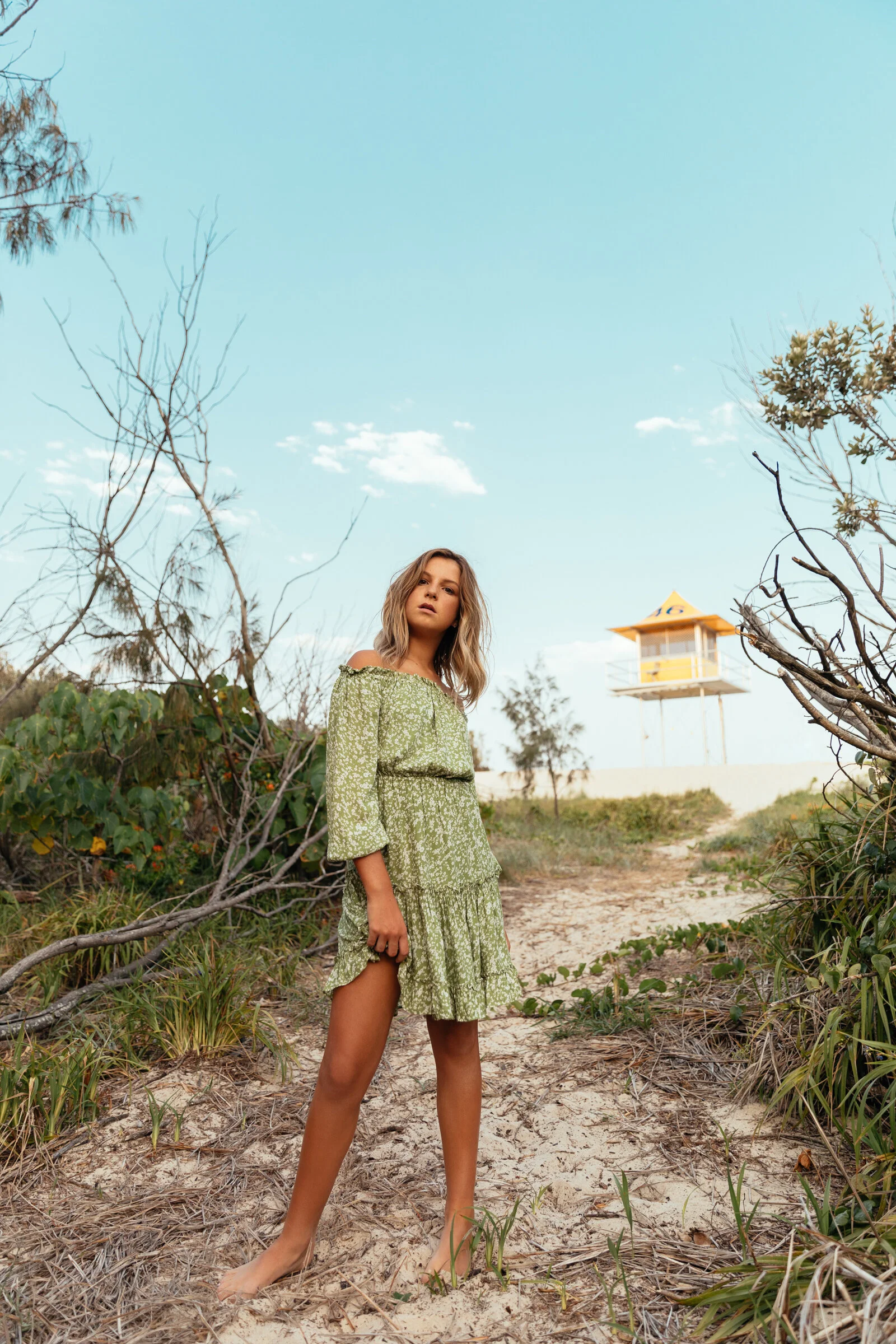 A young woman with long blond hair wearing a green dress, standing on a sandy path surrounded by sparse vegetation and leafless trees, under a clear blue sky with a lifeguard tower in the background.