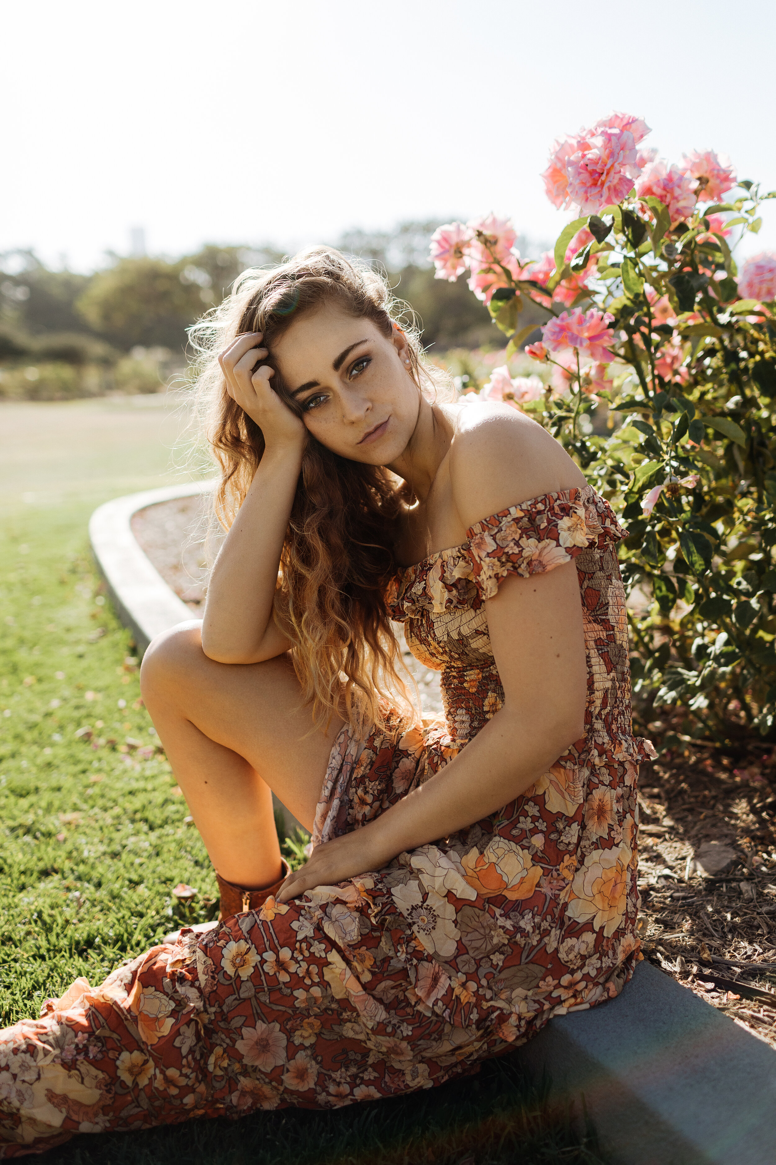 A young woman with red hair and blue eyes sitting on the grass near pink bushes, wearing a floral dress and brown boots, with one hand on her head and the other resting on her lap, outdoors on a sunny day.