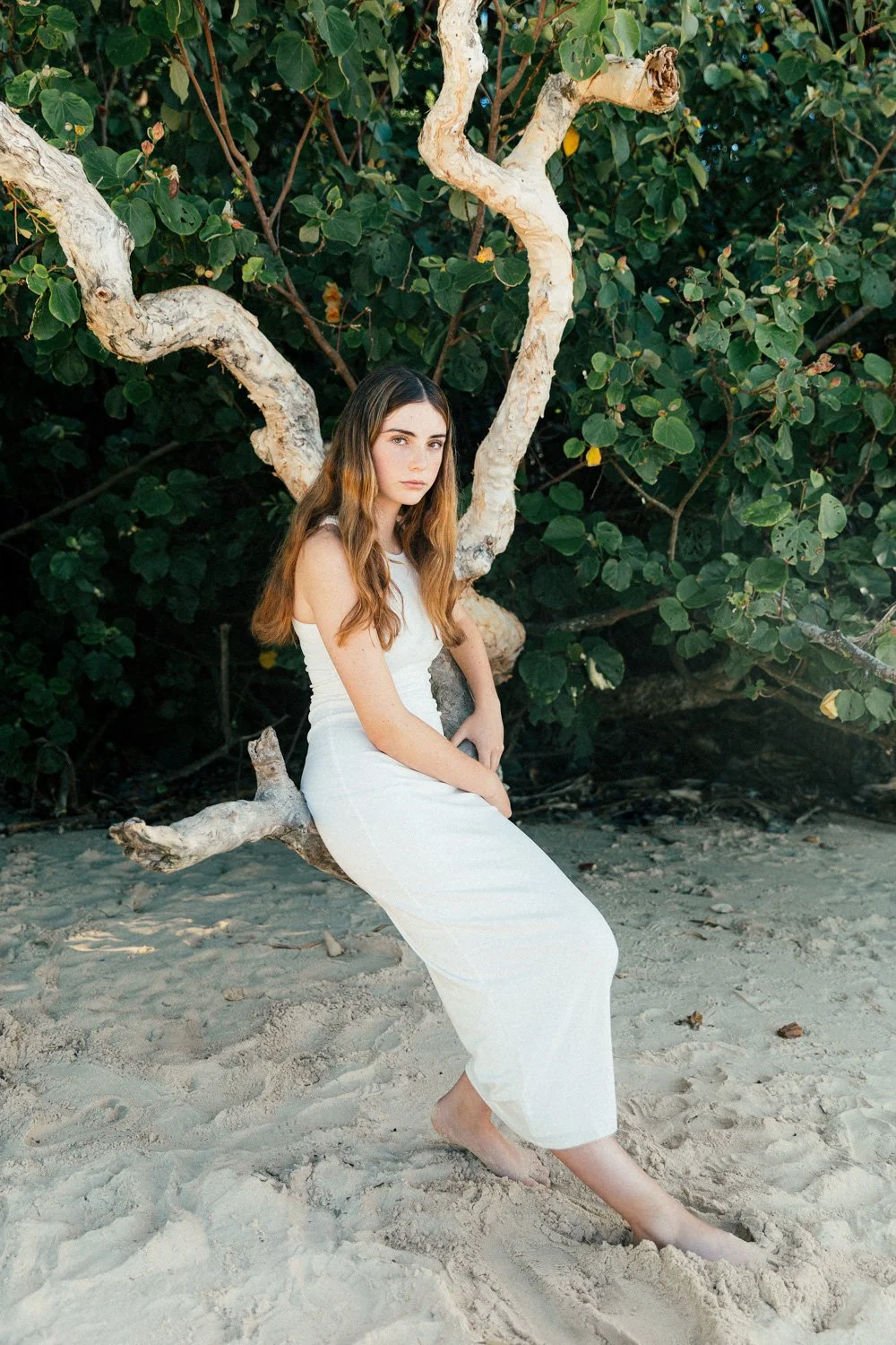 A young woman in a white dress sitting on a twisted driftwood branch on a beach with green foliage in the background.