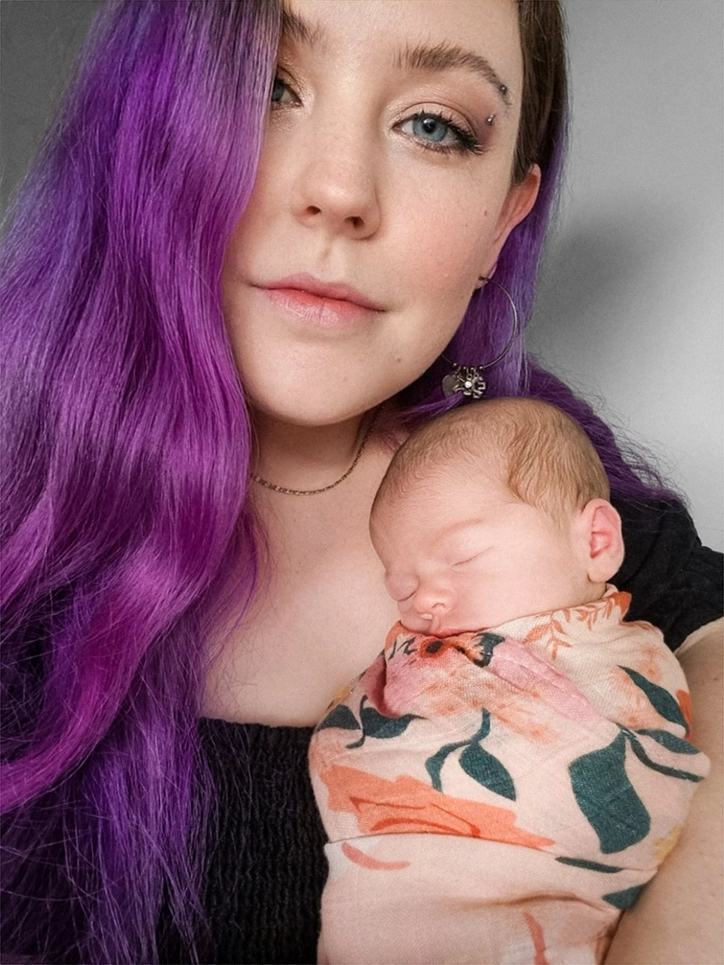 A young woman with long, wavy purple hair, fair skin, and distinctive eyebrow piercings is holding a sleeping infant wrapped in a floral-patterned blanket. The woman is wearing hoop earrings and has makeup that enhances her eyes.