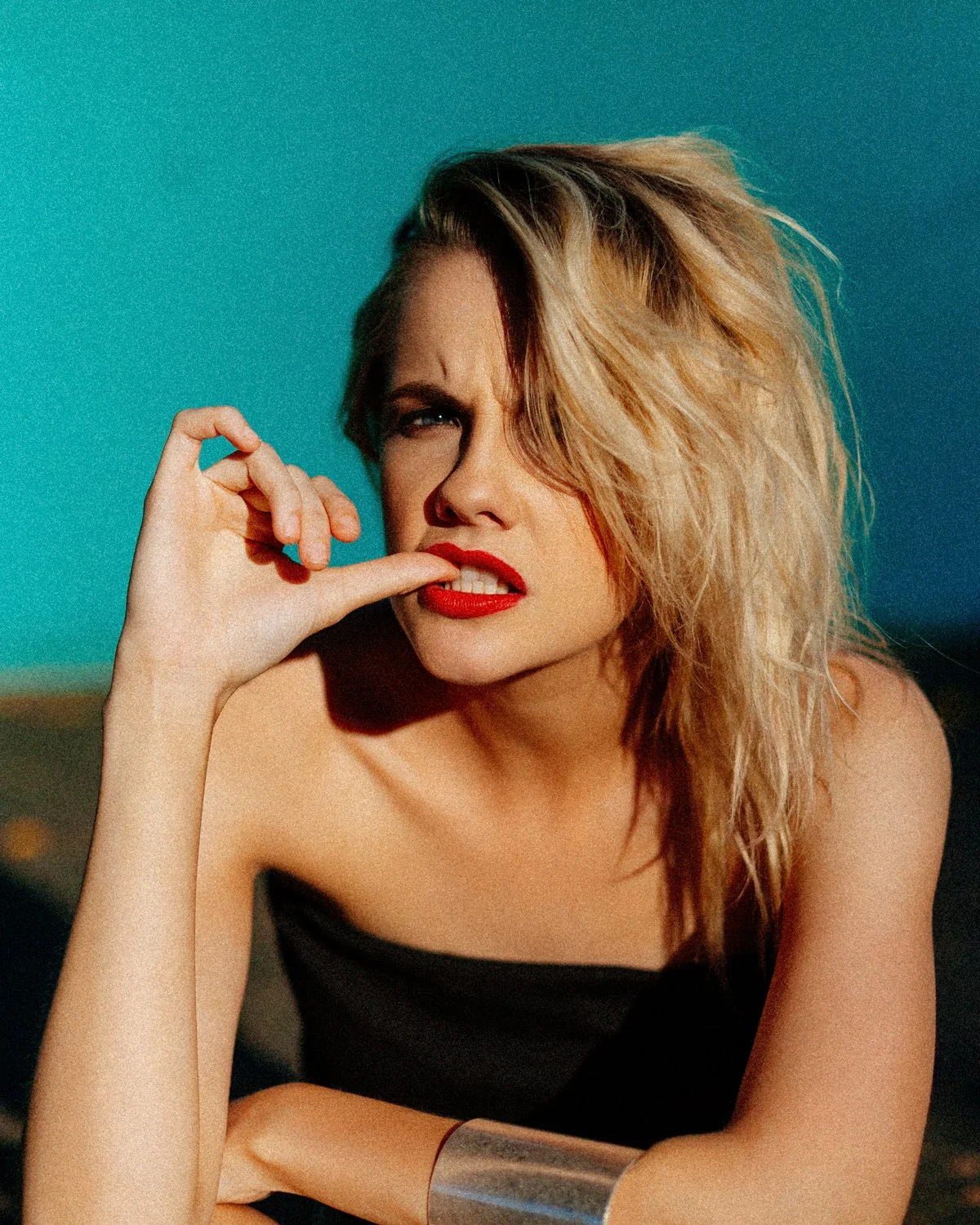 A woman with wavy blonde hair and red lipstick biting her finger against a blue background.