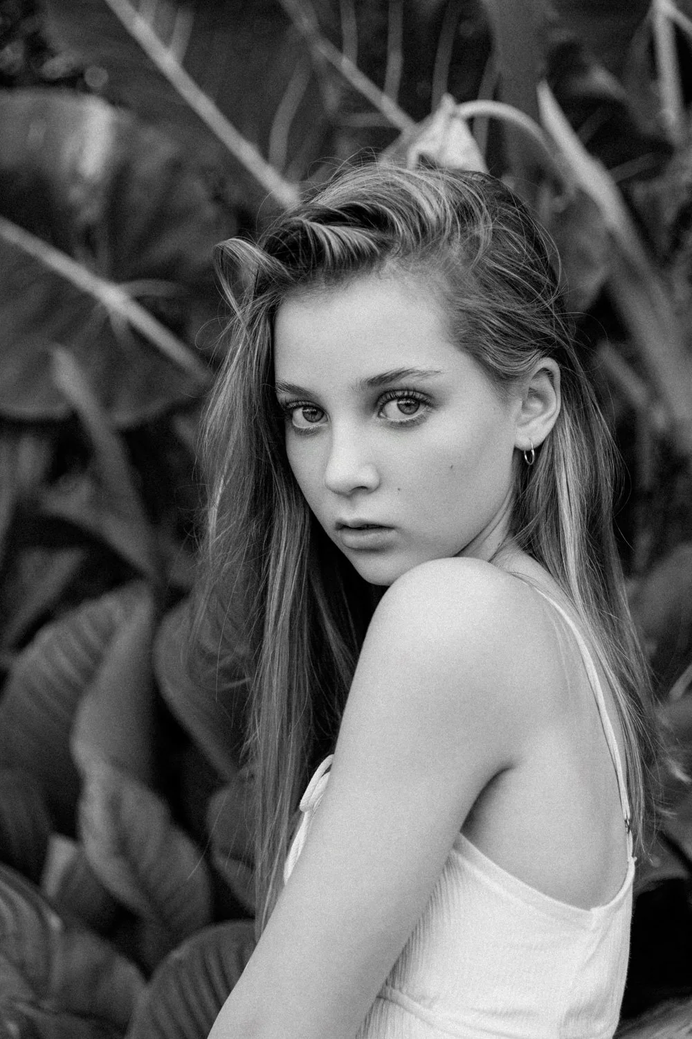 Black and white photo of a young woman with long hair, wearing a sleeveless top, looking over her shoulder at the camera, with a background of large leaves.