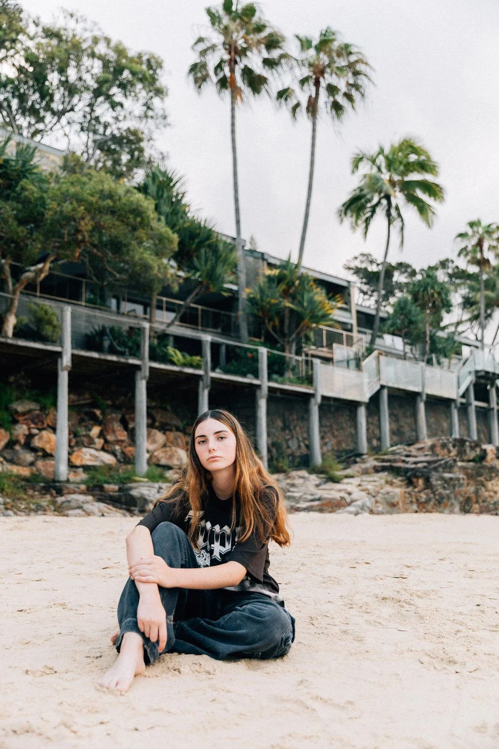 A young woman with long brown hair sitting on a sandy beach, wearing a black T-shirt and black pants, with houses and tall palm trees in the background under an overcast sky.