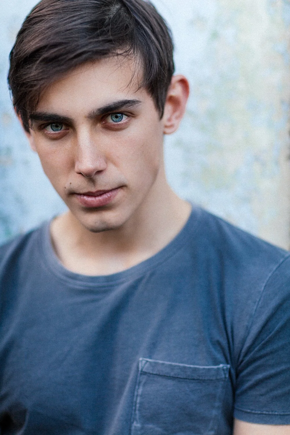 A young man with dark hair and blue eyes wearing a dark gray t-shirt looking directly at the camera. The background is a blurred, light-colored wall.