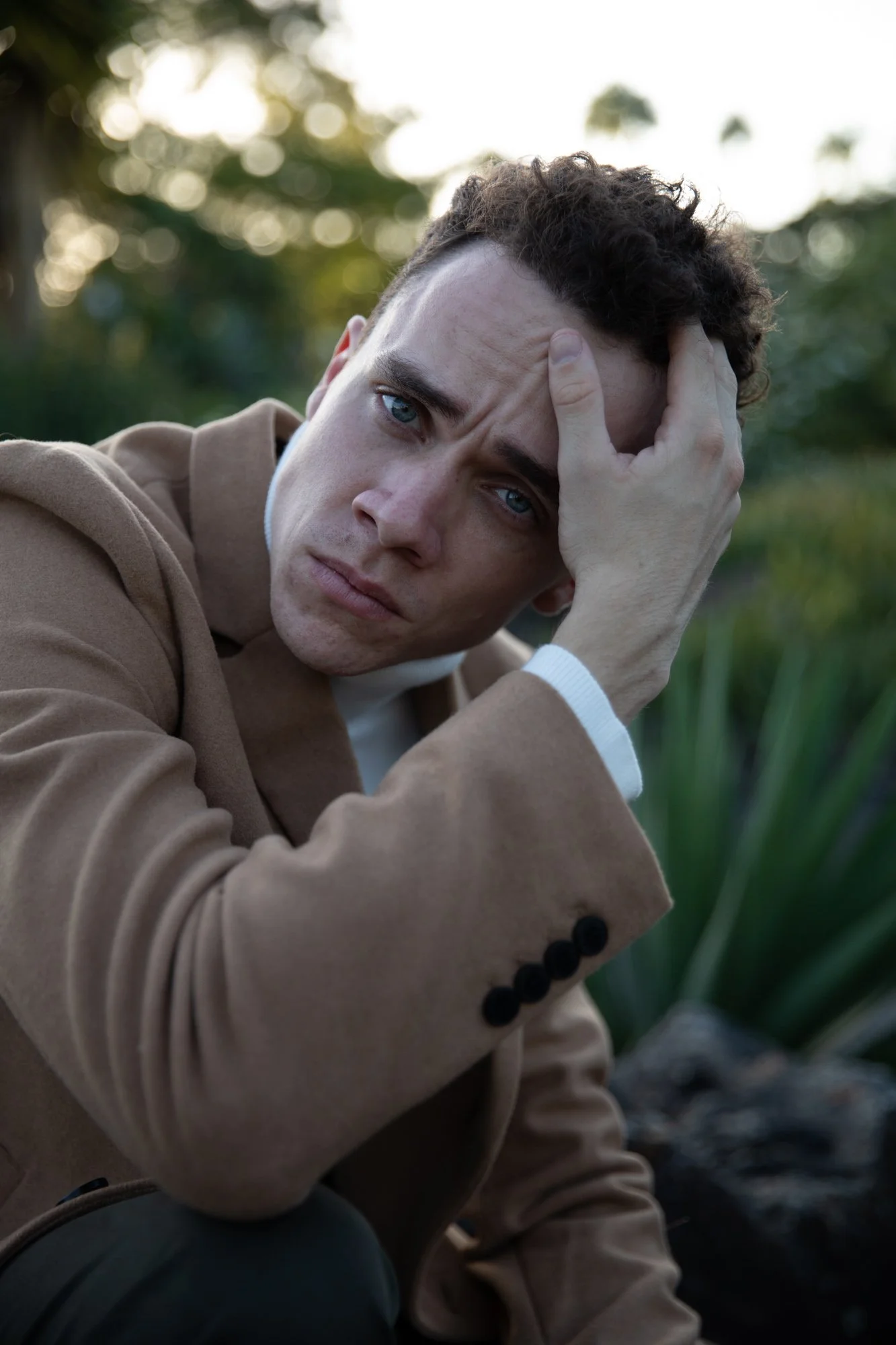 A young man with curly hair and blue eyes is sitting outdoors, resting his head on his hand, with a contemplative expression. He is wearing a tan blazer and a white shirt, with greenery and blurred sunlight in the background.
