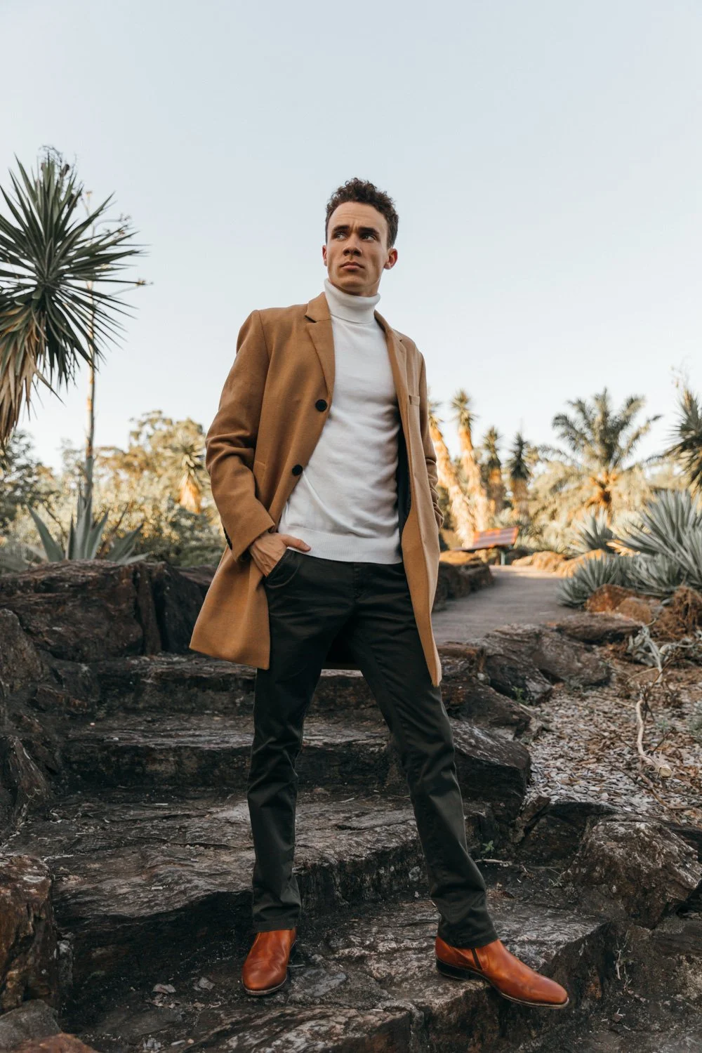 A man in a tan coat, white turtleneck, black pants, and brown boots standing outdoors on rocky steps. Desert plants and palm trees are in the background. The man has his hands in his pockets and looks off to the side.