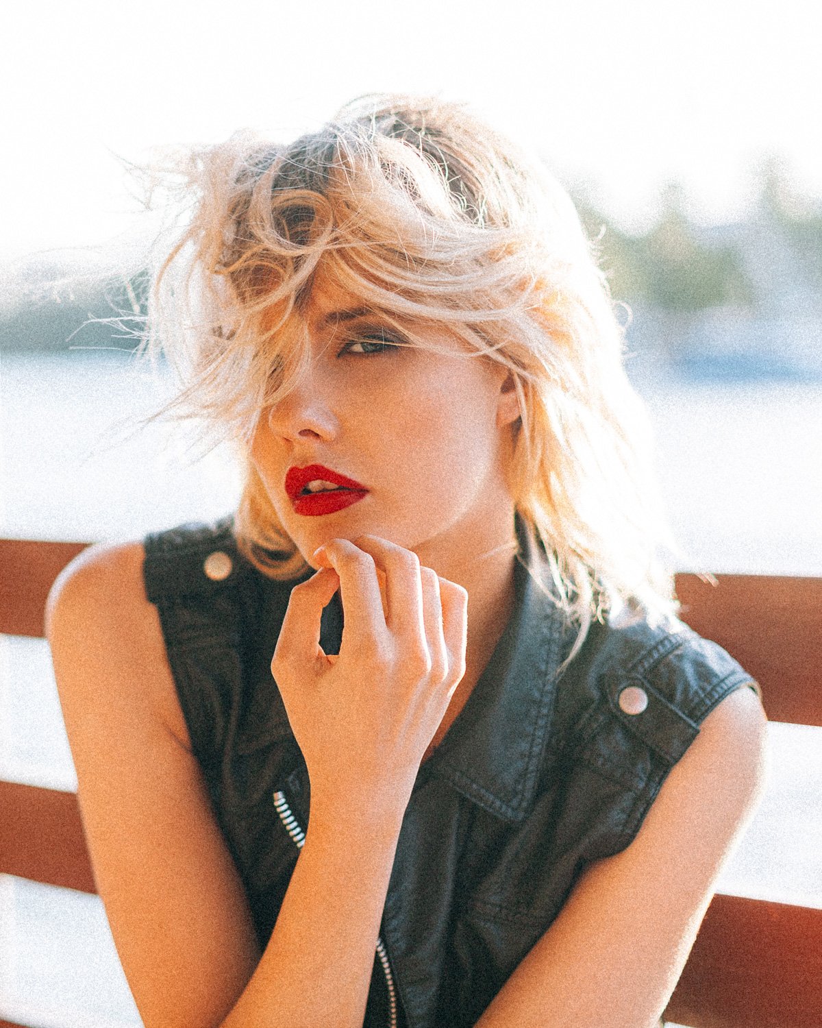 A young woman with short, wavy blonde hair and red lipstick, wearing a sleeveless black leather vest, sitting outdoors with sunlight highlighting her face.