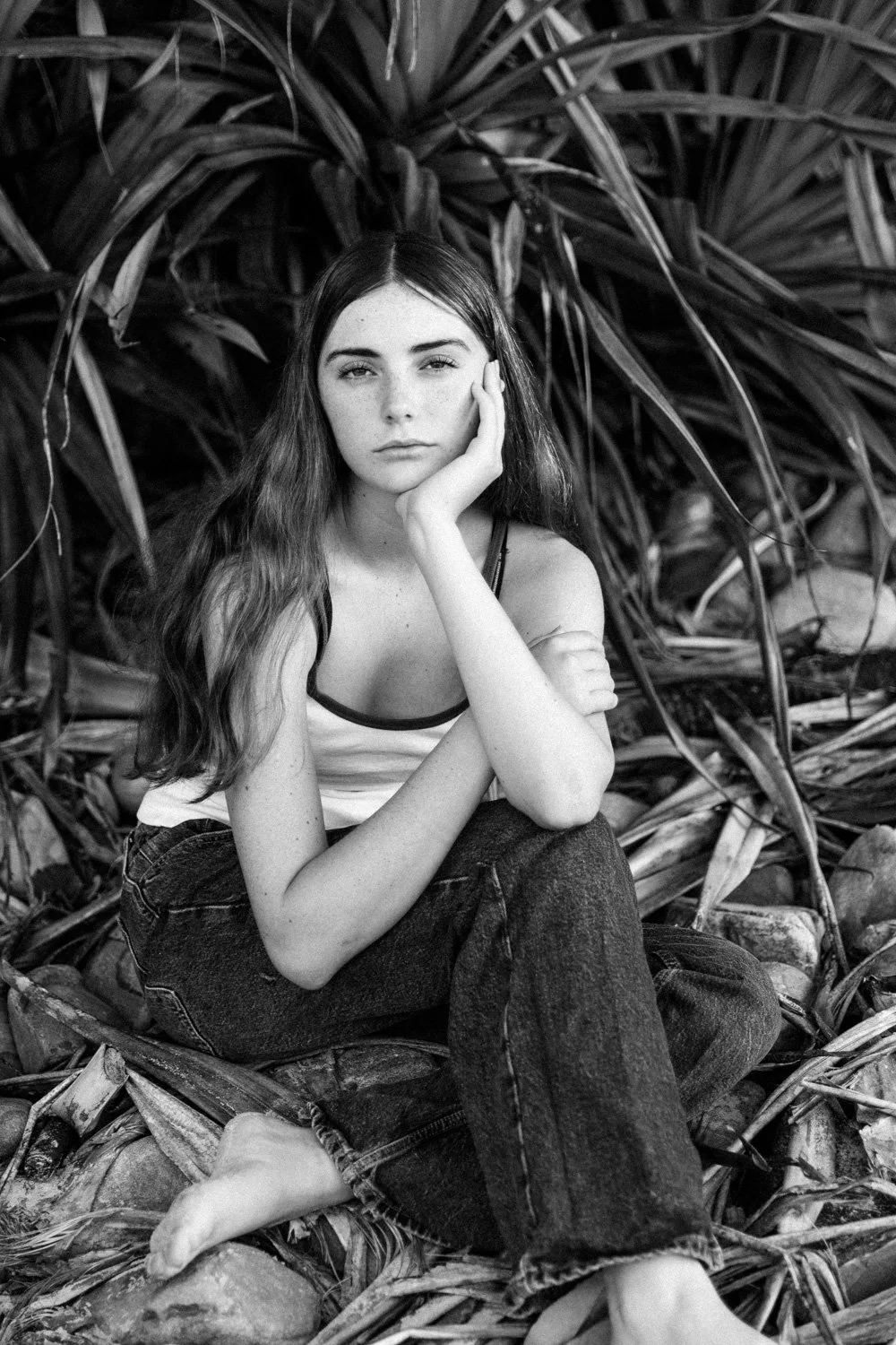 Black and white photo of a young woman with long wavy hair, sitting among large leaves or plants, resting her head on her hand, looking at the camera with a neutral expression.