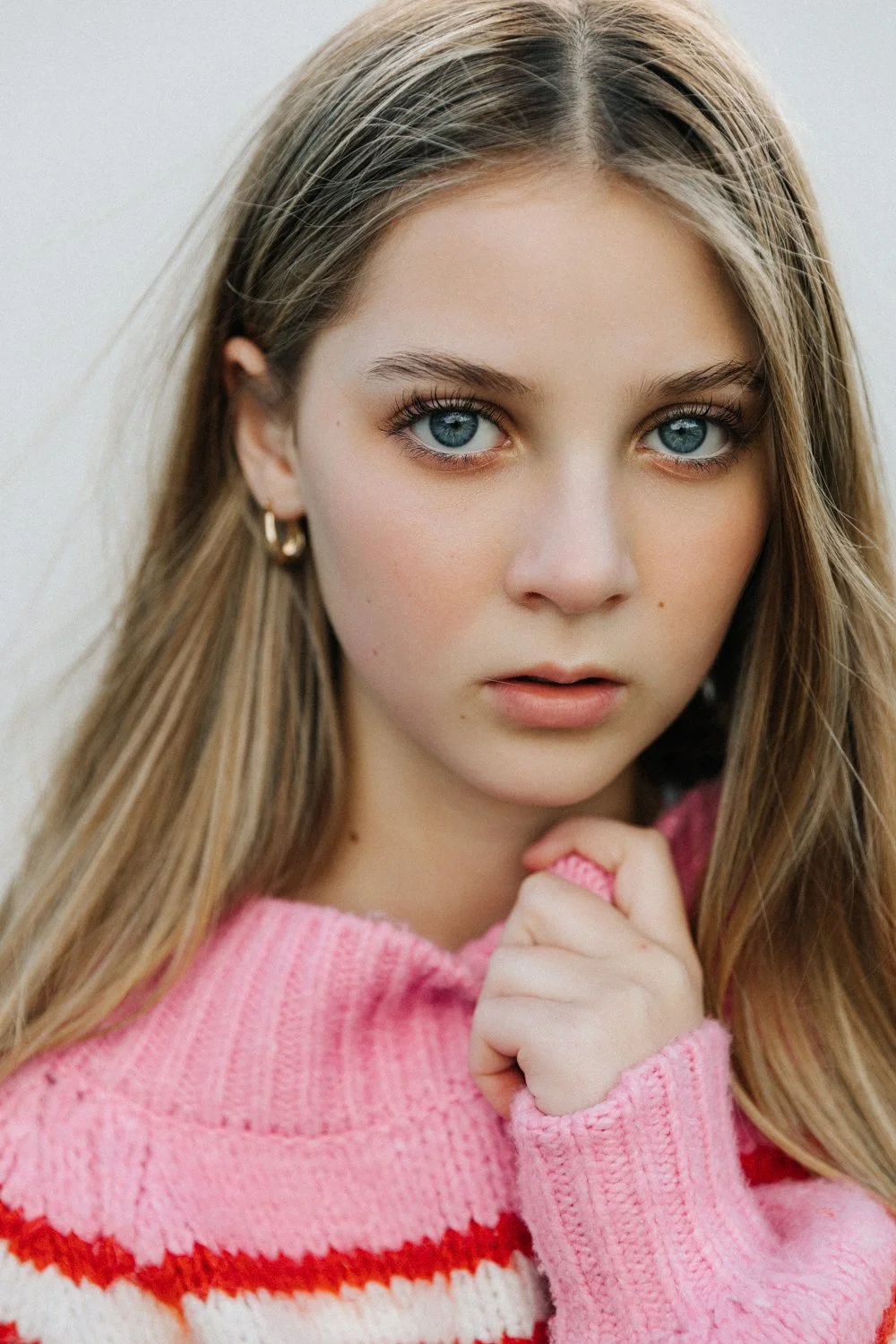 Close-up of a young girl with blue eyes and long light brown hair, wearing a pink and red striped sweater, resting her chin on her hand, looking serious.
