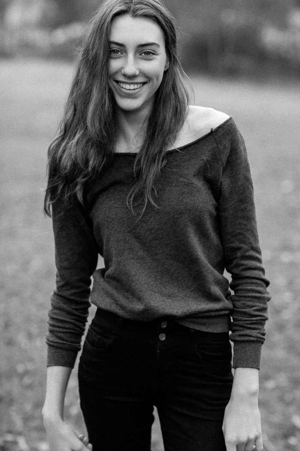 Black and white photo of a young woman smiling, standing outdoors with a blurred natural background, wearing a dark casual long sleeve shirt and dark pants.