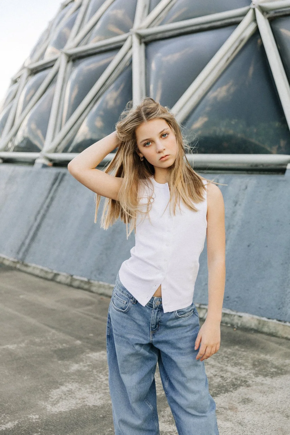 A young woman with blonde hair in a white sleeveless top and blue jeans, standing outdoors against a modern glass and metal building, with her left hand resting on her head and gazing at the camera.