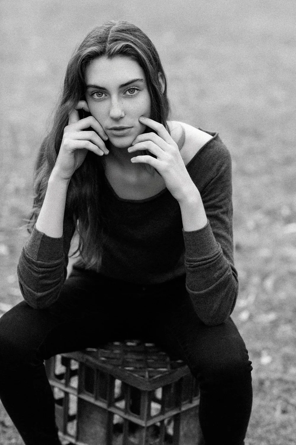 A young woman with long hair sitting on a crate outdoors, looking directly at the camera with her chin resting on her hand.