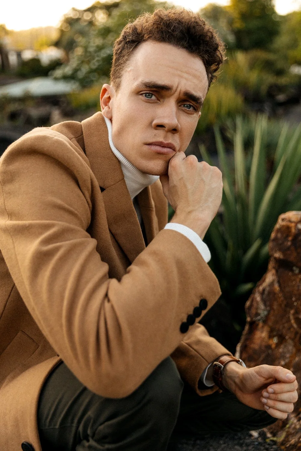 A young man with curly brown hair, blue eyes, wearing a tan coat and white turtleneck, sitting outdoors near plants and rocks, resting his chin on his hand and looking contemplative.