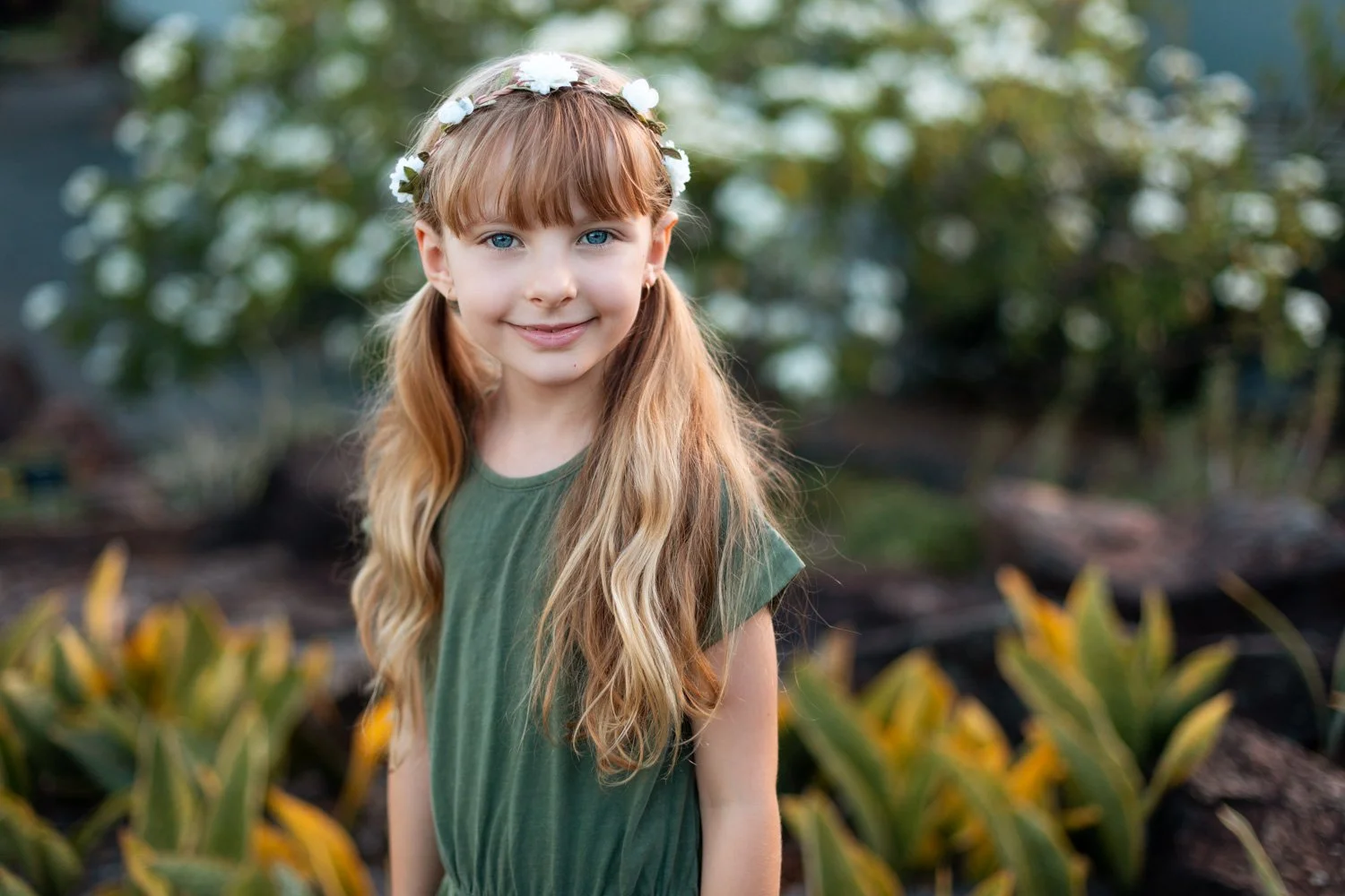 A young girl with long, wavy blonde hair and blue eyes, wearing a green shirt and a white floral headband, smiling outdoors in a garden with plants and trees in the background.