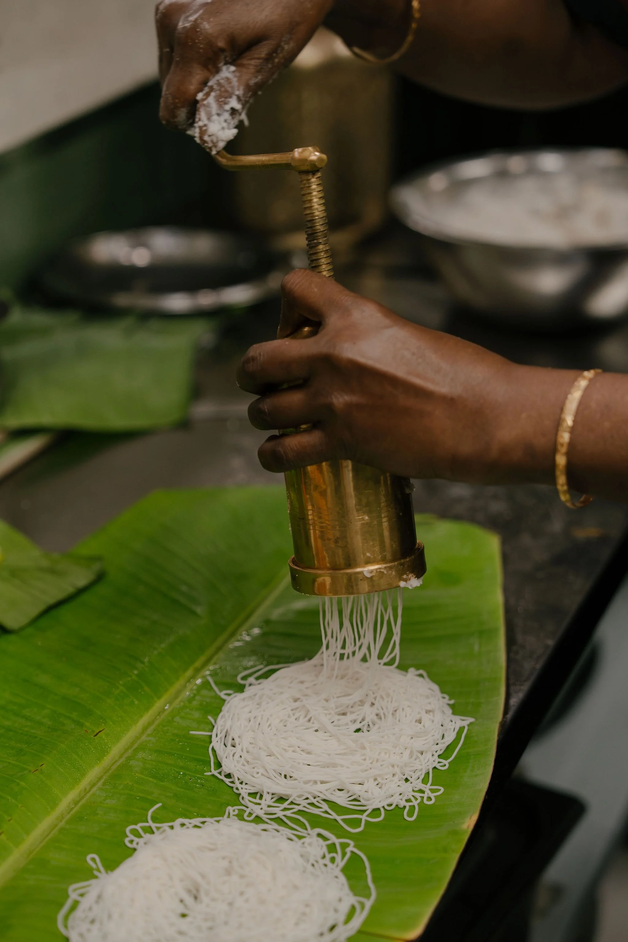 Person pressing a noodle press onto a green banana leaf, with freshly made noodle strands hanging down, in a kitchen setting.