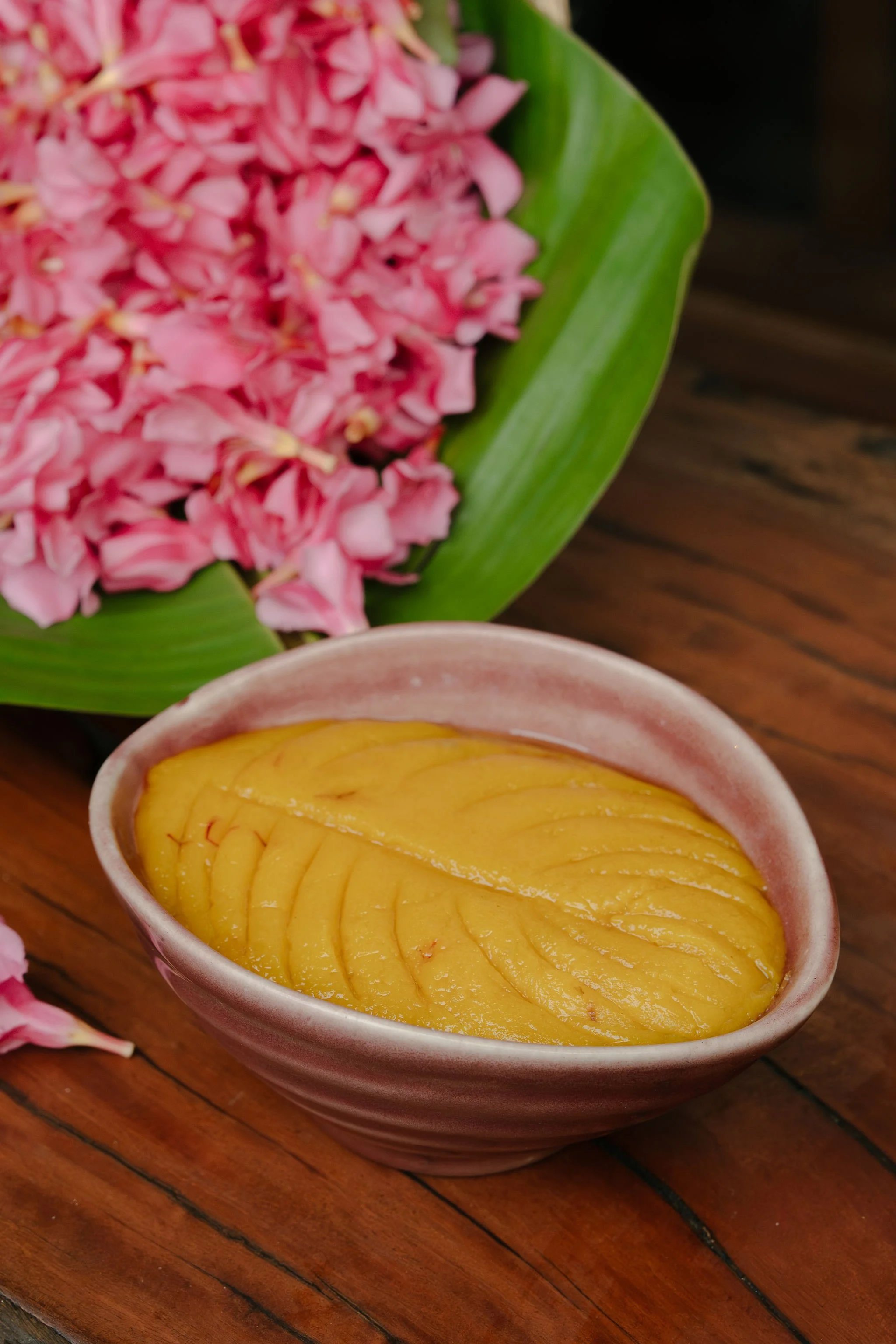Badam halwa in a ceramic bowl on a wooden surface, pink flowers with green leaves in the background.