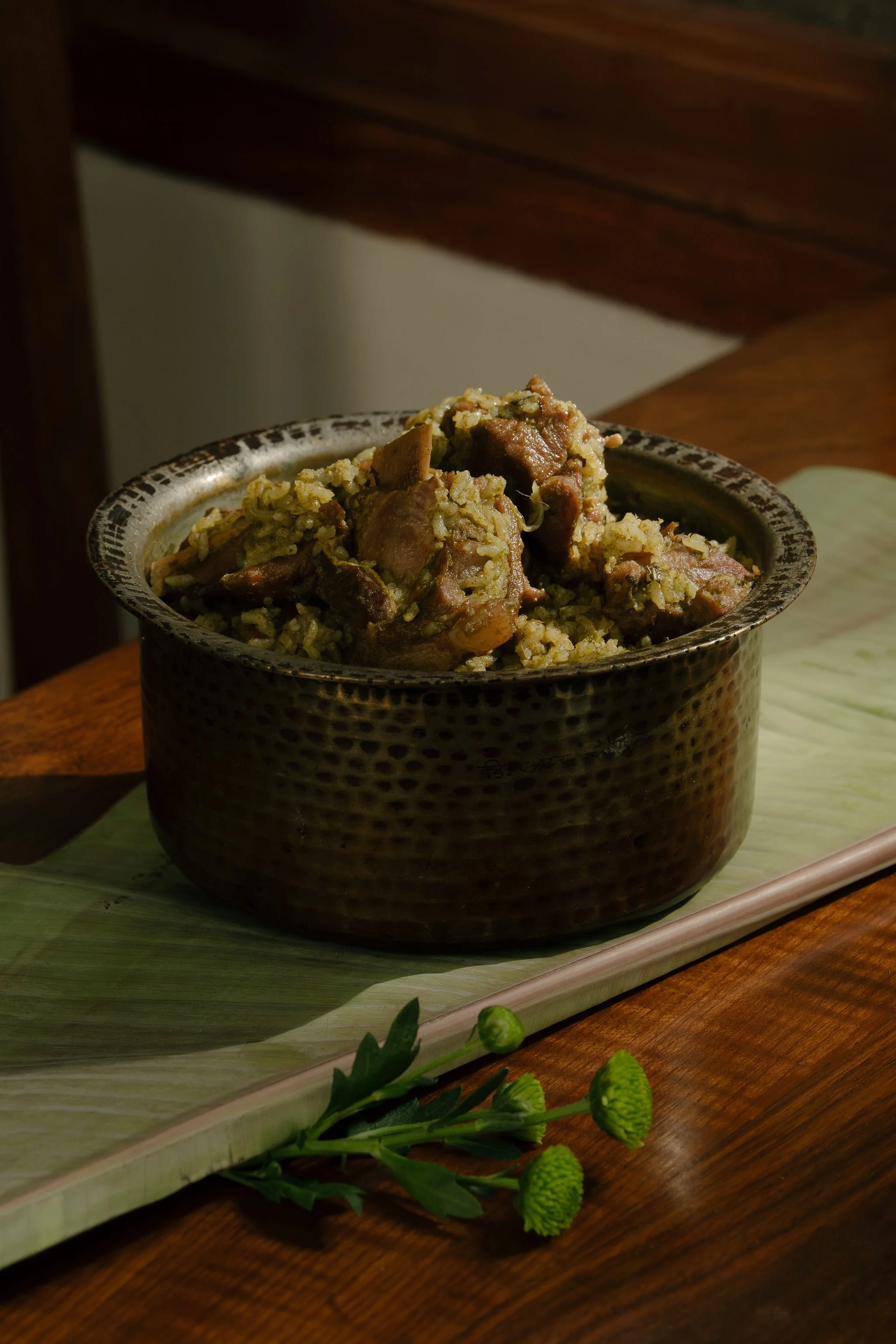 A bowl of rice and meat stew on a wooden table with a banana leaf underneath and green flowers in front.