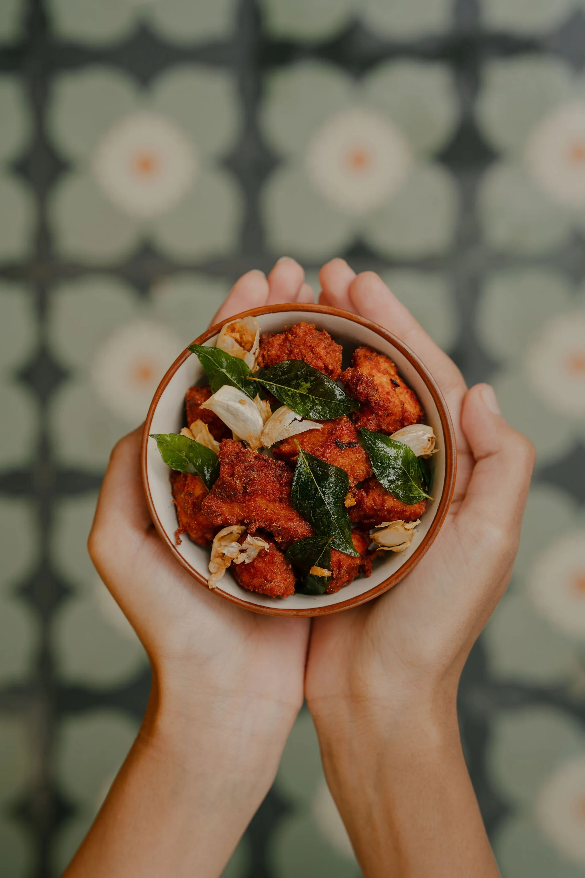 Person holding a bowl of fried chicken garnished with bay leaves and garlic cloves, against a patterned background.