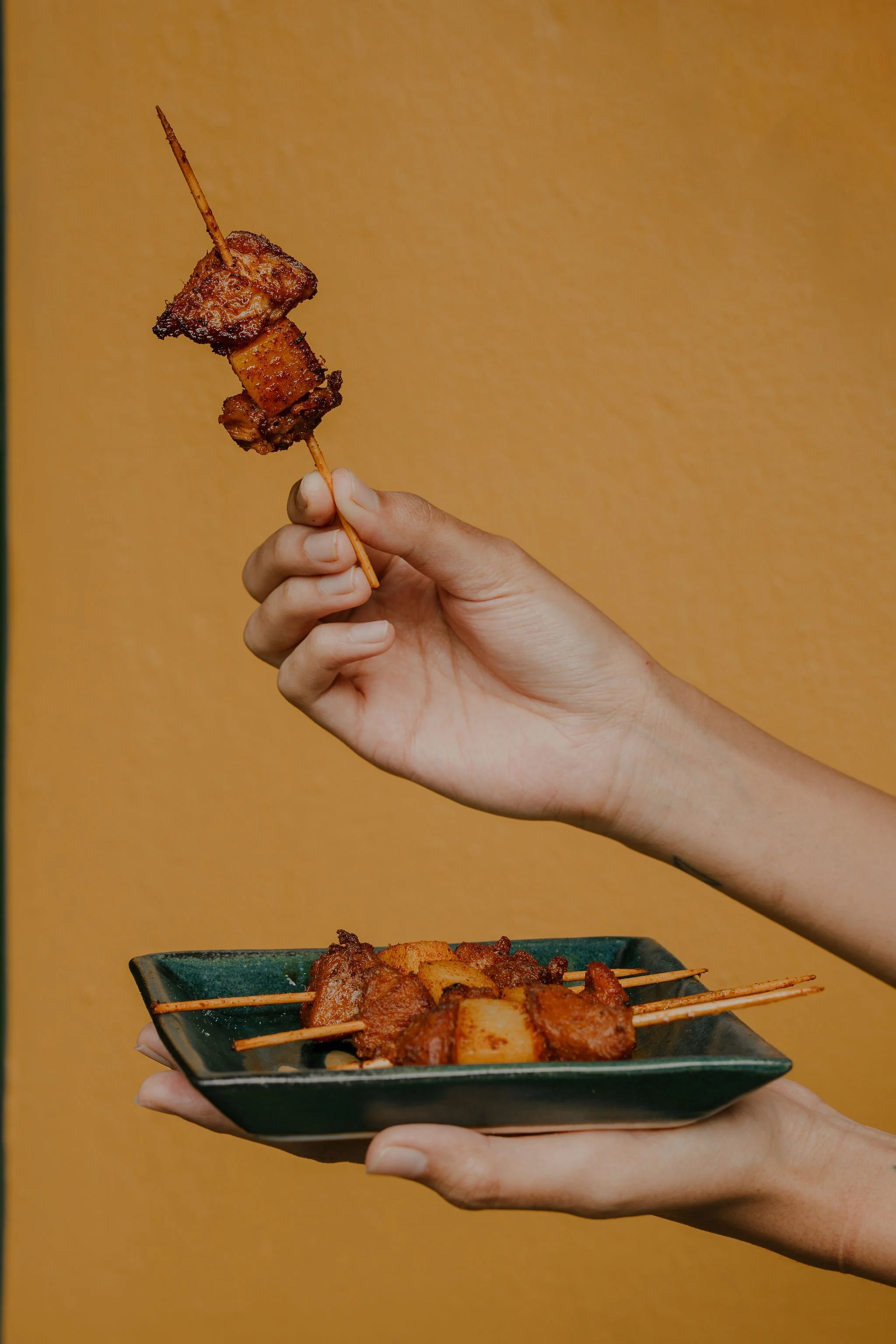 Person holding a skewer of grilled meat and pineapple over a green plate with more skewers of grilled food against a yellow background.