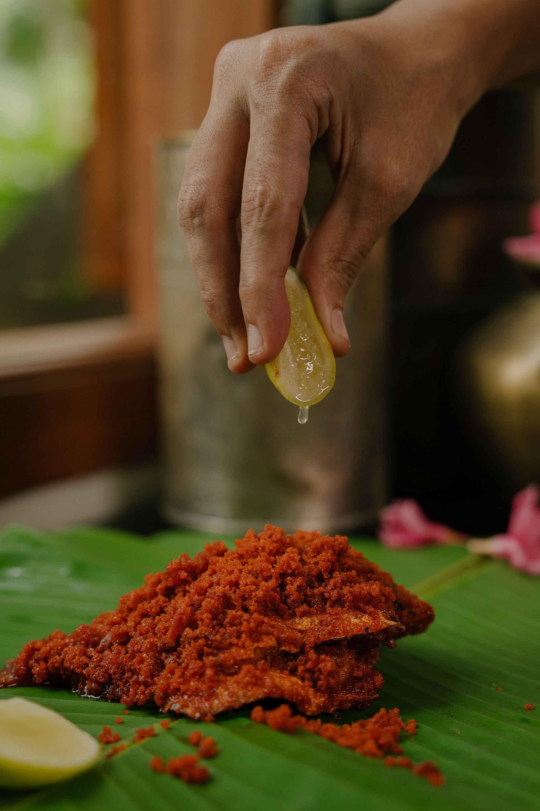 Hand squeezing lemon over a fried fish fillet on a banana leaf, with a lime wedge in the foreground.