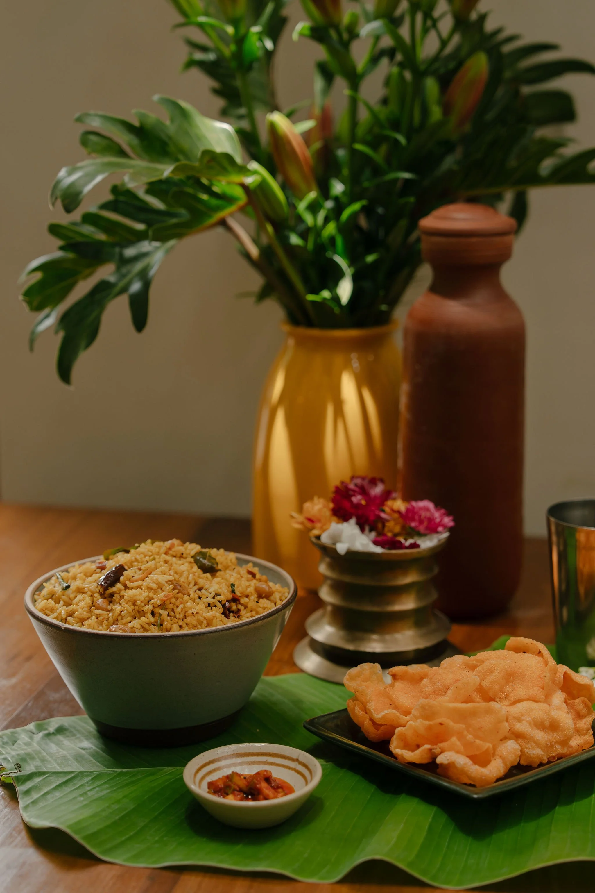 A bowl of rice with vegetables, a small dish of pickles, crispy fried snacks on a black plate, and a banana leaf on a wooden table with two vases and flowers in the background.