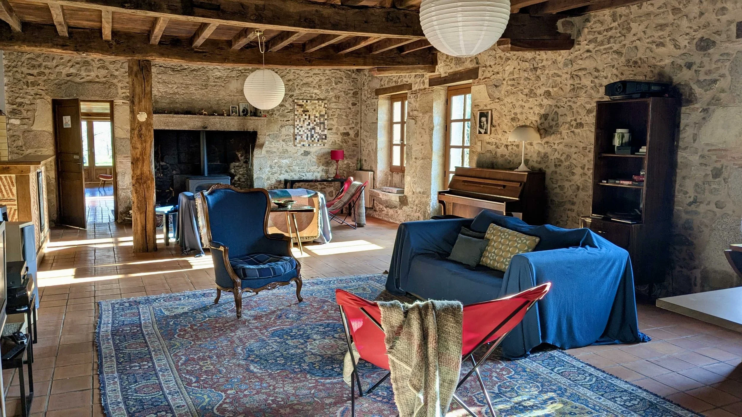 Salon chaleureux avec mobilier vintage, cheminée en pierre, meubles en bois, tapis traditionnel, et murs en pierre, éclairé par la lumière naturelle.